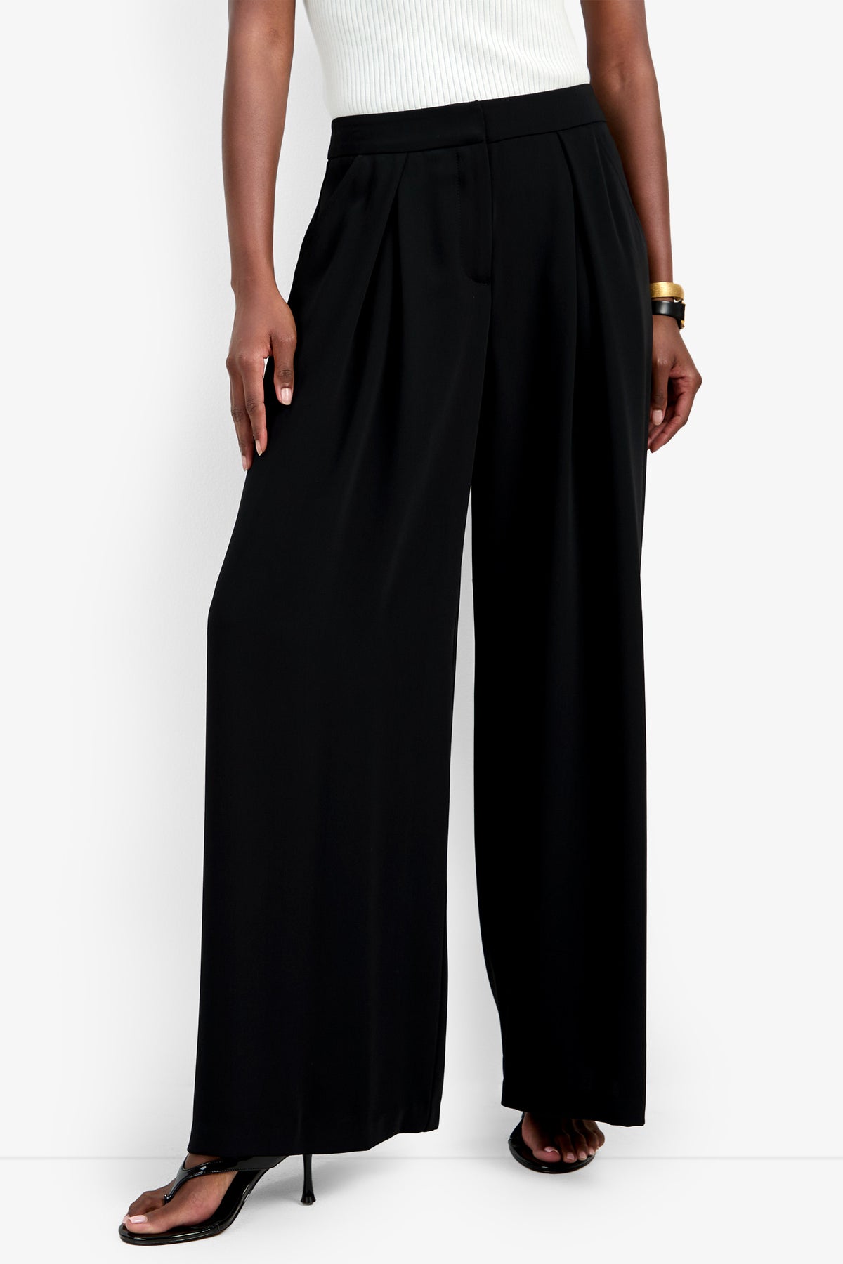 A person wearing a white sleeveless top, the black Saige Pant, black high-heeled sandals, and a gold bangle stands against a white background.