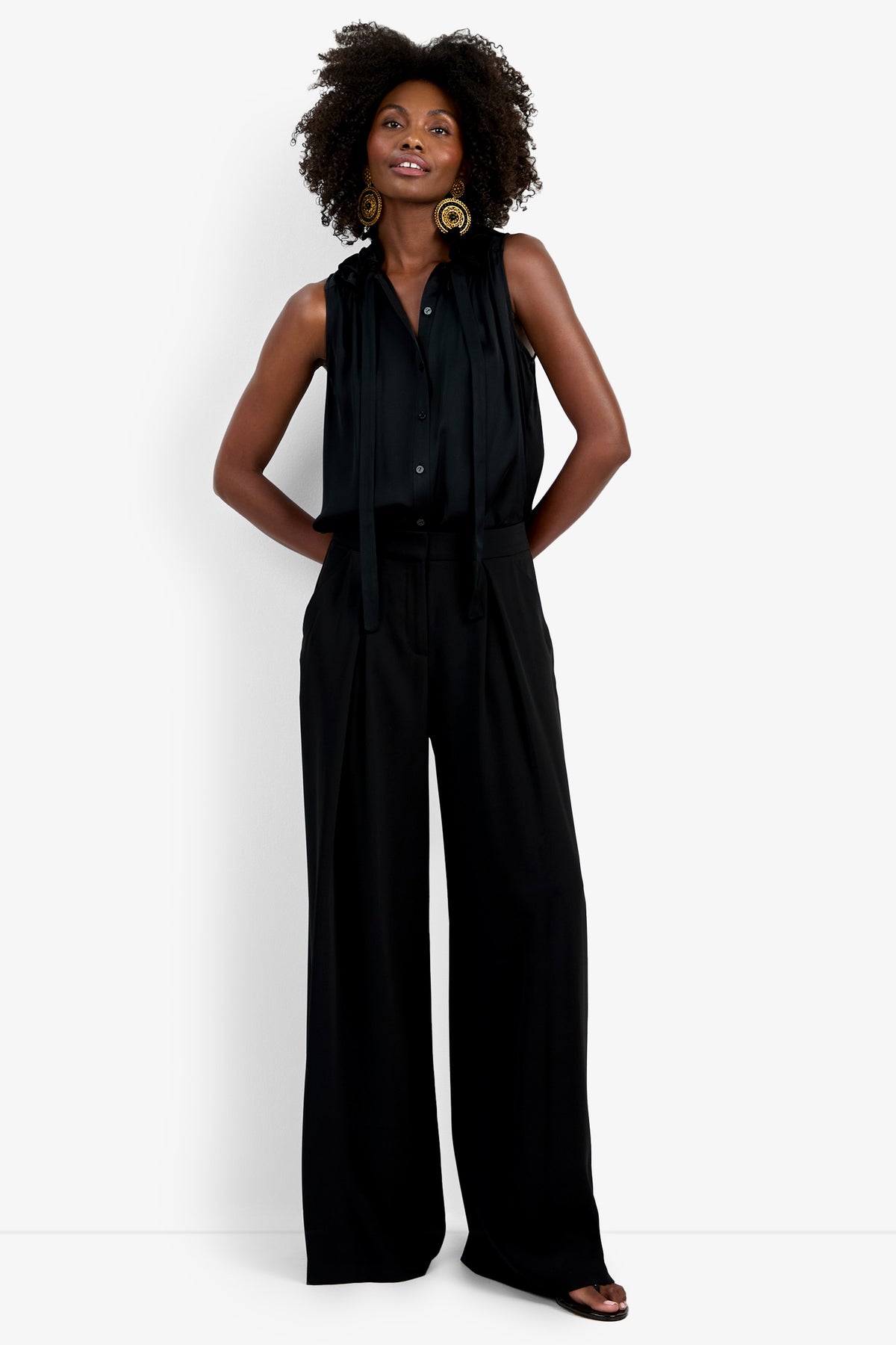 A woman stands confidently in the Saige Long Pant—wide-leg black trousers—paired with a sleeveless black blouse, gold statement earrings, and open-toed black shoes, against a plain white background.