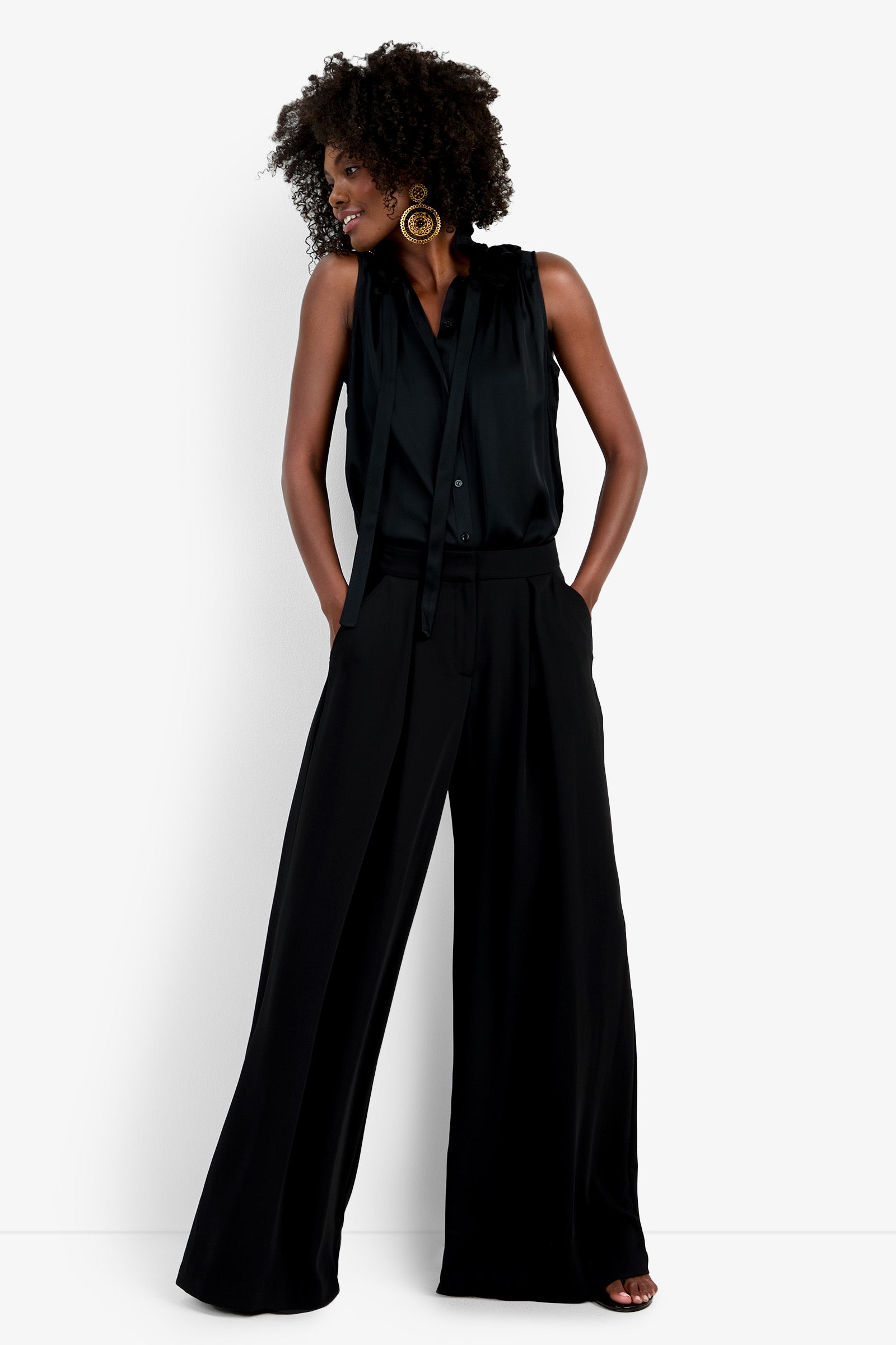 A woman with curly hair wears wide-leg black Saige Long Pants and a sleeveless black blouse with a tie detail. She stands against a plain white background, hands in pockets, looking to the side.