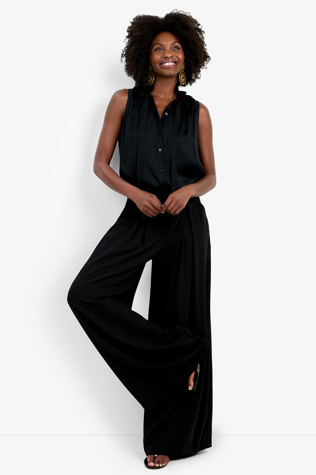 A woman with curly hair smiles against a white background, wearing the Saige Long Pant, a sleeveless black blouse, large round earrings, and black sandals.