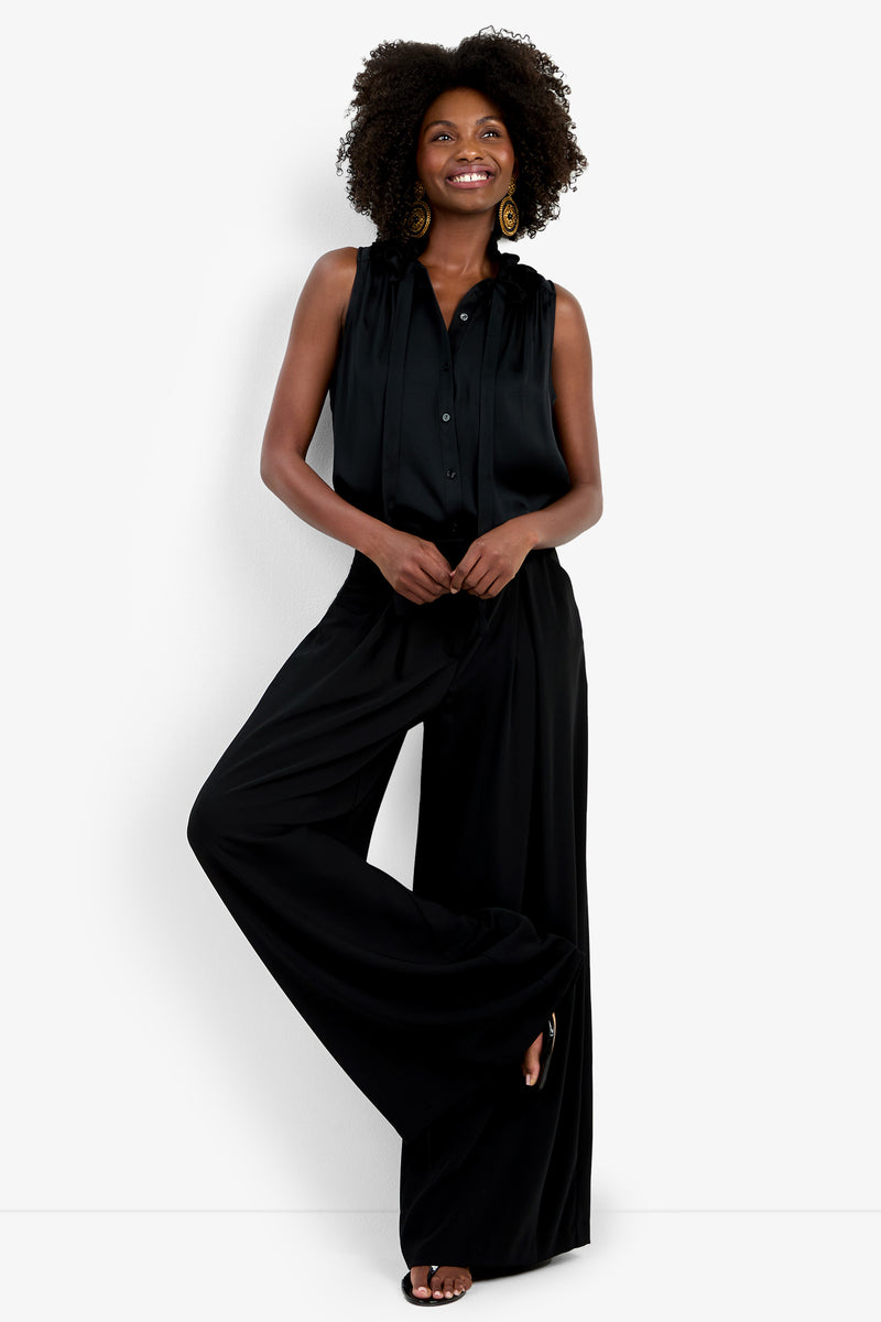 A woman with curly hair smiles against a white background, wearing the Saige Long Pant, a sleeveless black blouse, large round earrings, and black sandals.