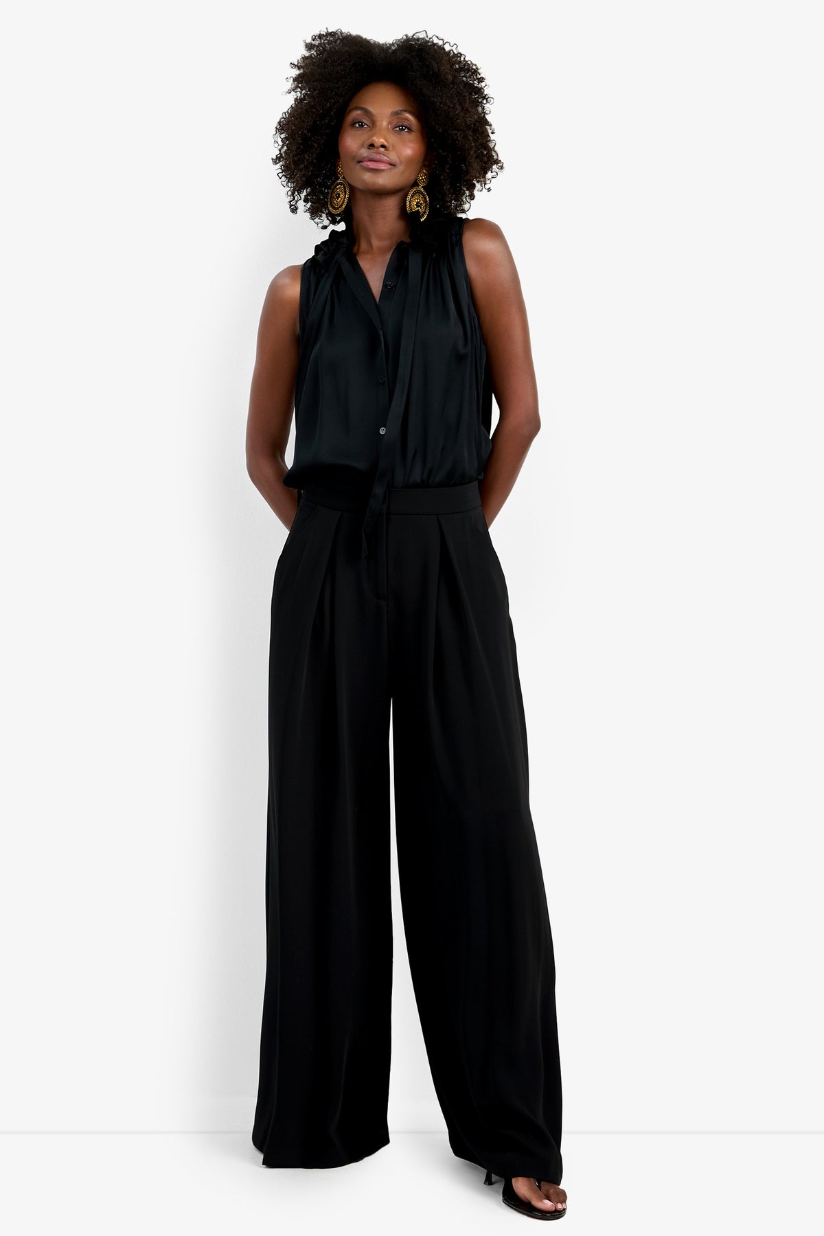 A woman with curly hair and gold statement earrings stands confidently against a white background, wearing the Saige Long Pant—a wide-leg black trouser—paired with a sleeveless black ruffled-collar blouse and black open-toe shoes.