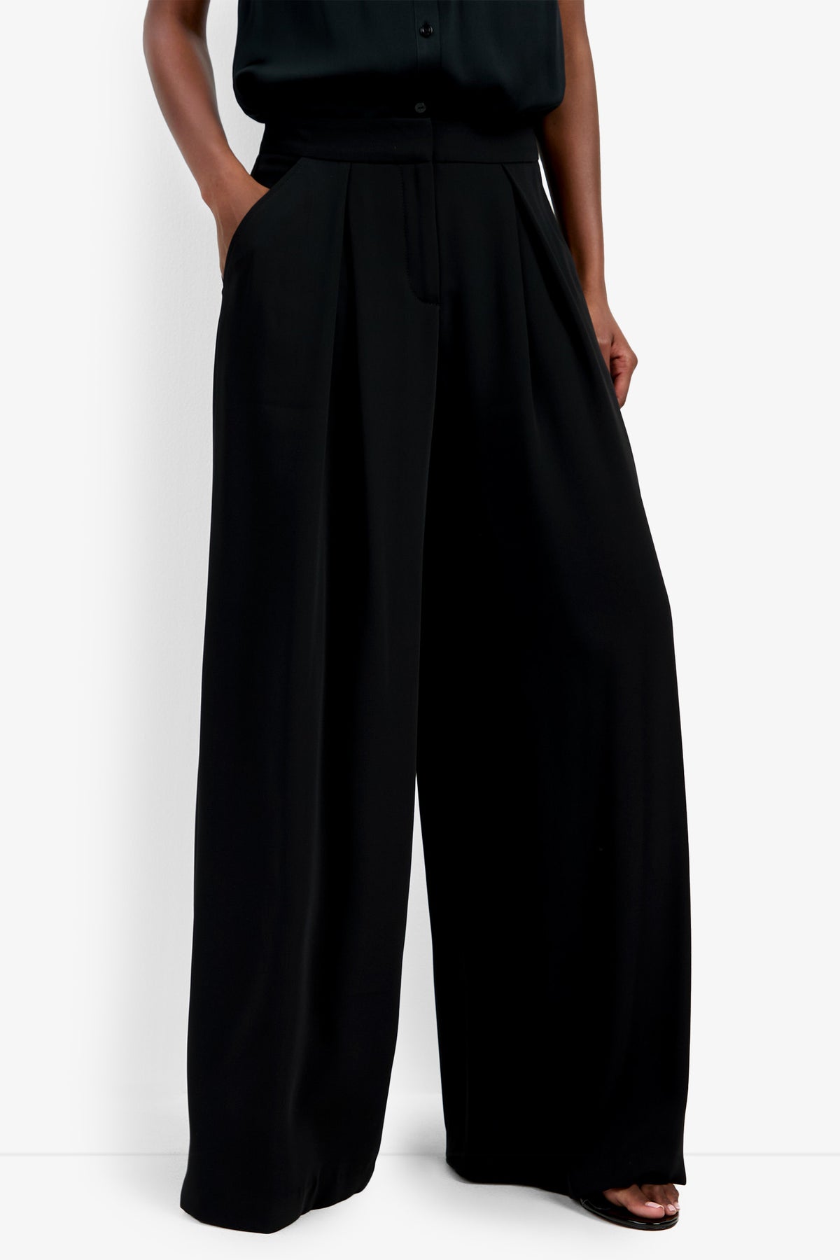 Model wears the Saige Long Pant—high-waisted, wide-leg black trousers with pockets—paired with a black top. The look features a loose, flowing silhouette for an elegant, modern style.