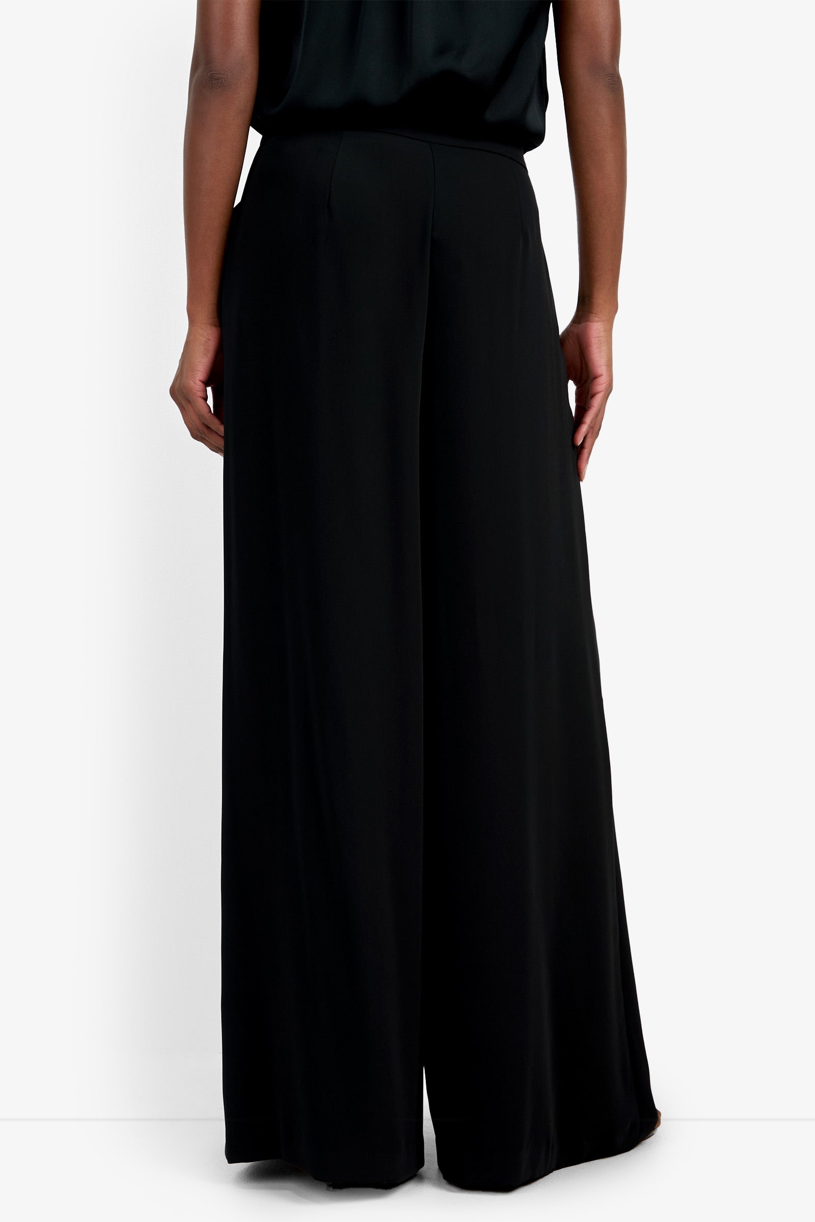 A person stands facing away against a white background, wearing loose-fitting, high-waisted Saige Long Pant in black and a matching black top.
