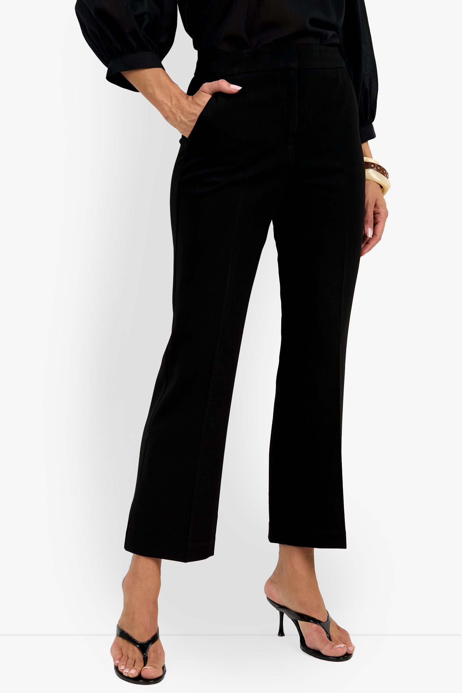 A woman wears the Allisan Pant, paired with a black blouse with voluminous sleeves, black heeled sandals, and gold bracelets, standing against a white background with one hand in her pocket.
