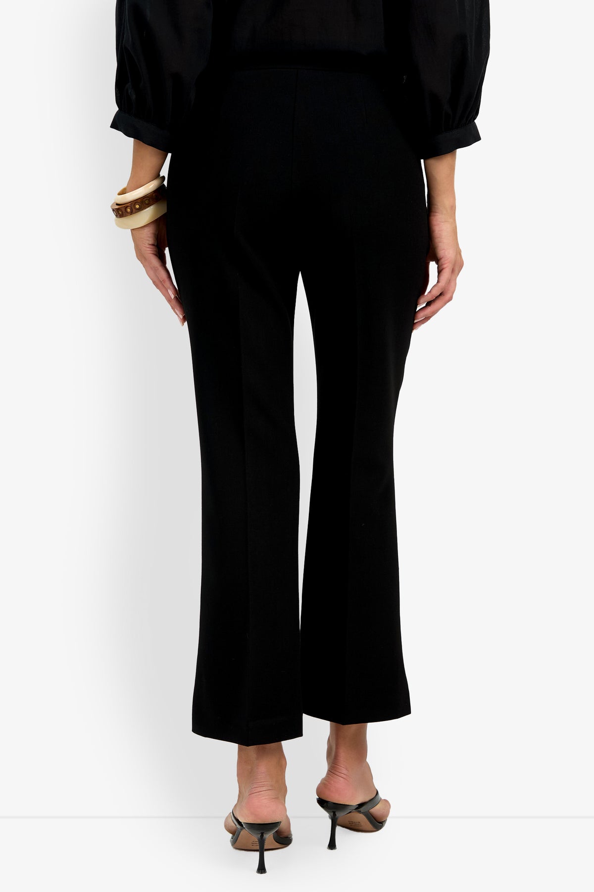 A person stands facing away on a plain white background, wearing the Allisan Pant in black, a black long-sleeve blouse, heeled sandals, and bracelets on their left wrist.