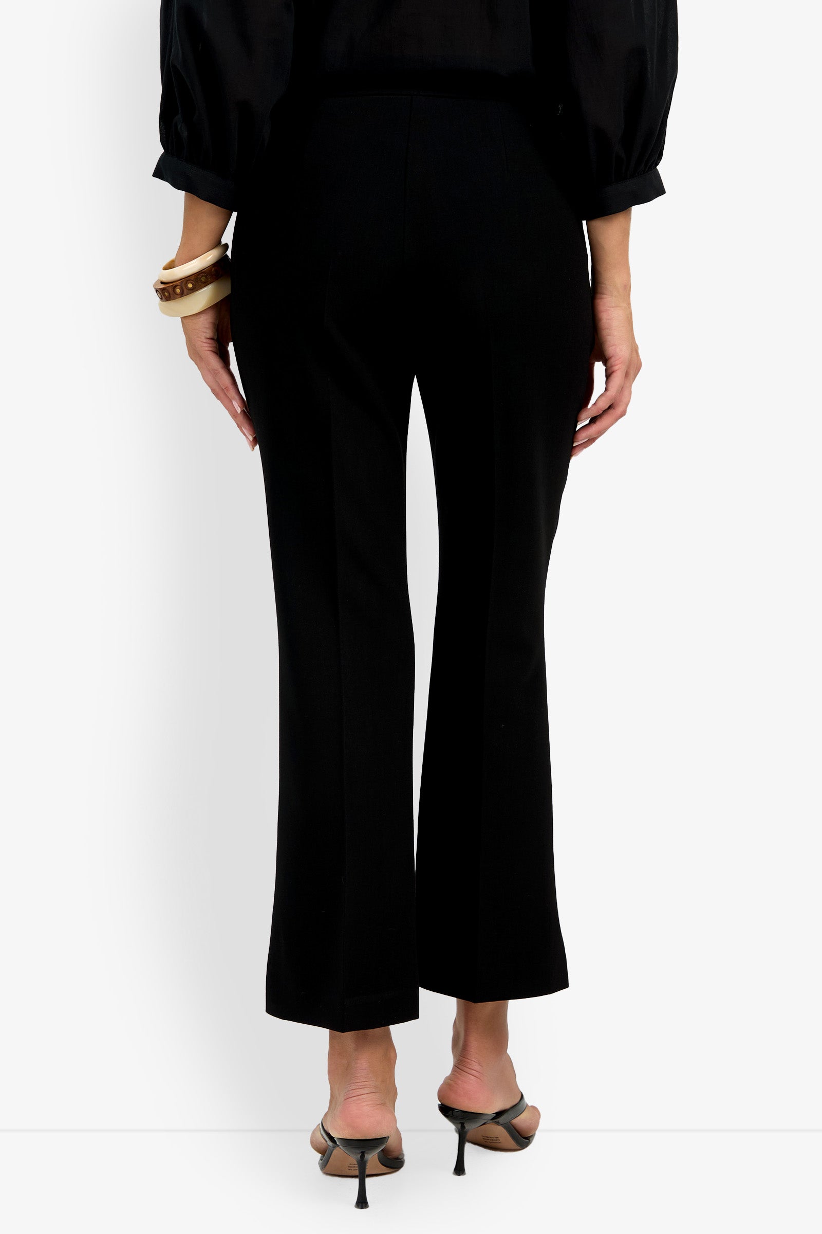 A person stands facing away on a plain white background, wearing the Allisan Pant in black, a black long-sleeve blouse, heeled sandals, and bracelets on their left wrist.