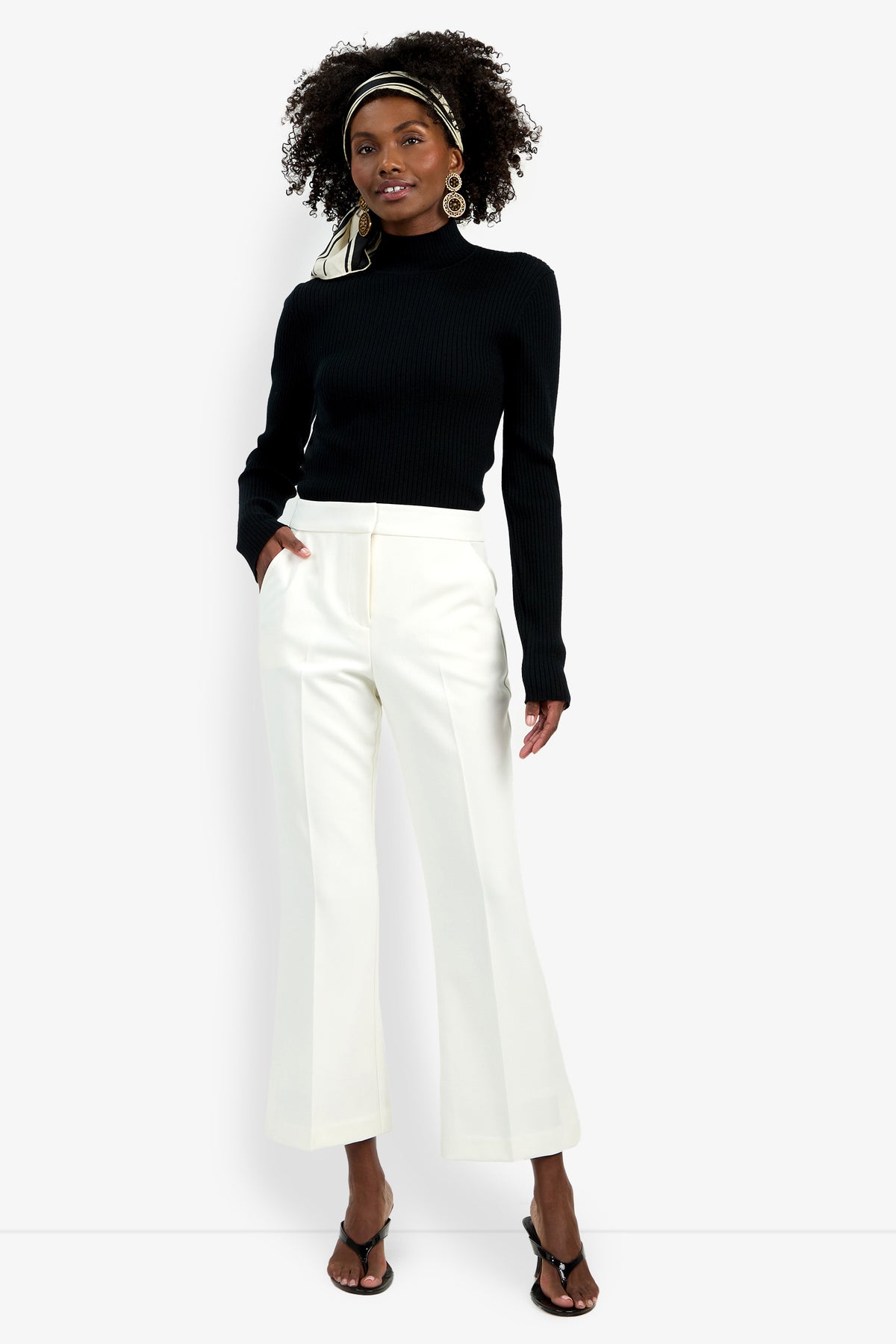 A woman stands against a white background, smiling slightly with one hand in her pocket. She wears the Allisan Pant in white, paired with a black long-sleeve top, black heeled sandals, hoop earrings, and a headscarf.