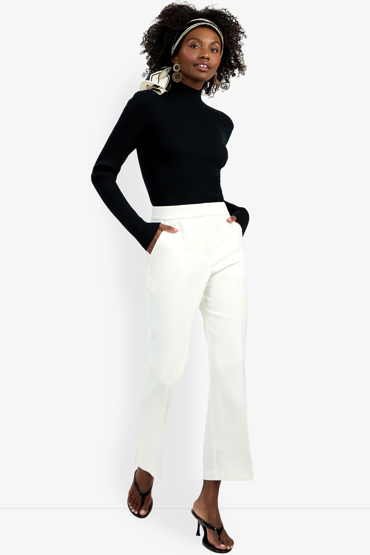 A woman stands confidently facing the camera, hands in her pockets, wearing the Allisan Pant in white with a black turtleneck sweater, black heeled sandals, large hoop earrings, and a patterned headscarf.