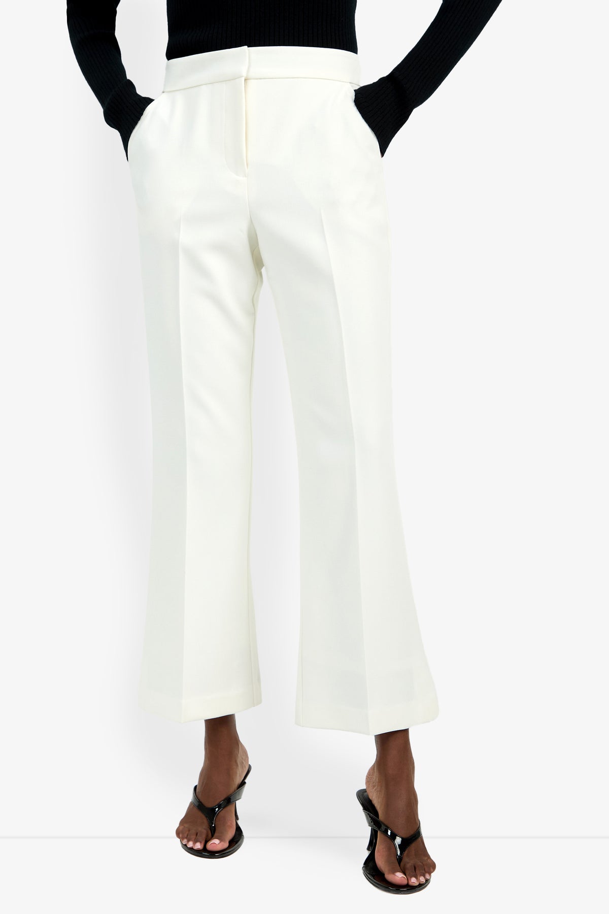 A person wears the Allisan Pant—white, high-waisted, wide-leg trousers—with hands in pockets, paired with a black long-sleeve top and black heeled sandals, standing against a plain white background.