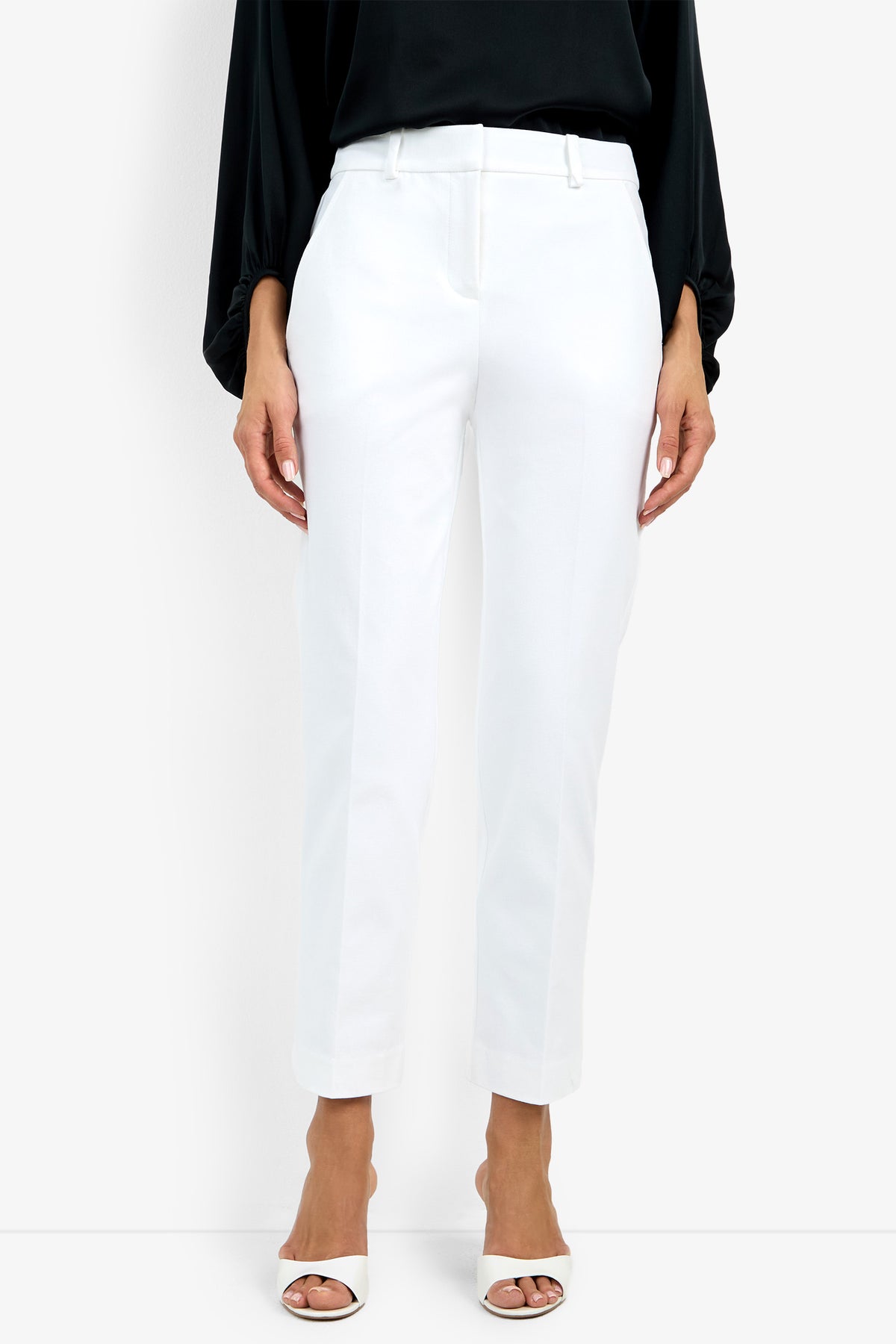 A person is shown from the neck down wearing the Audrey Pant, a white ankle-length trouser, with a black long-sleeve top and white open-toe heels against a plain white background.