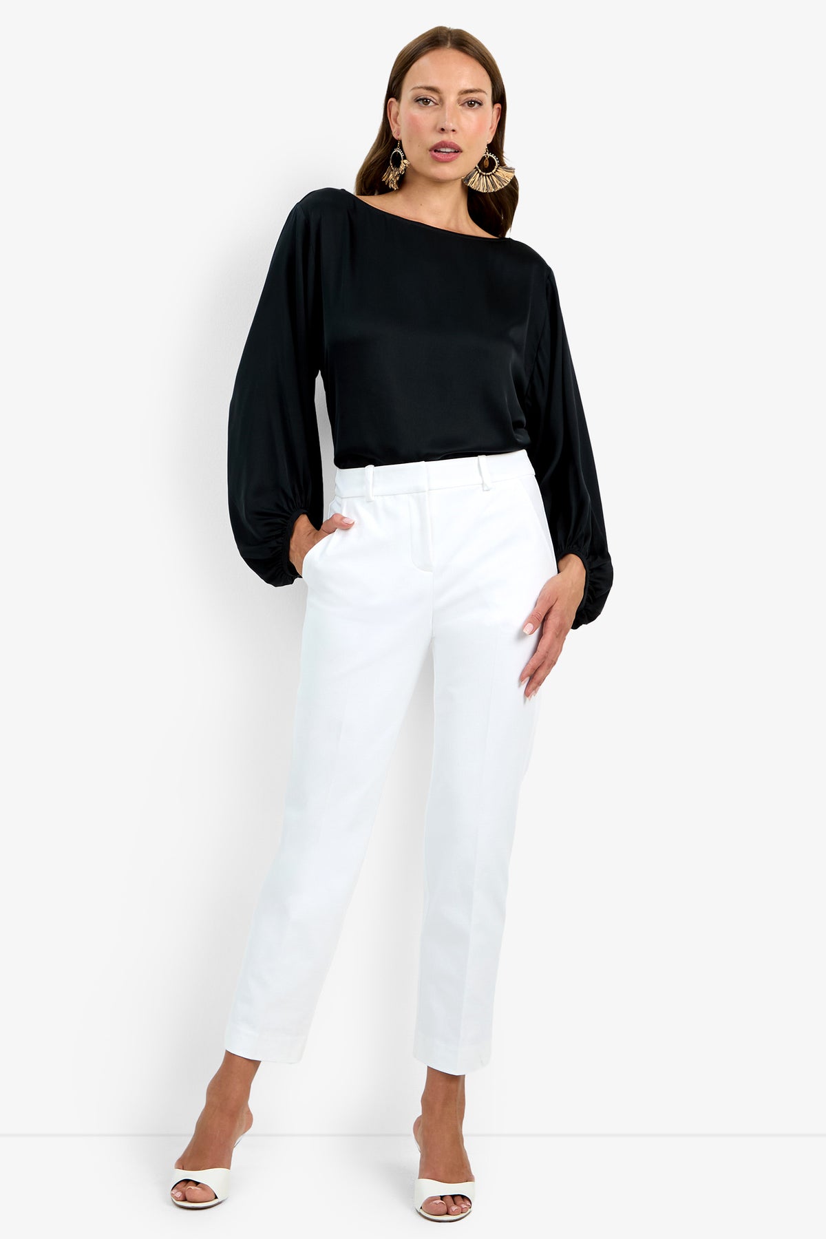 A woman with straight hair and a confident pose wears the Audrey Pant, high-waisted white trousers, paired with a black balloon-sleeve blouse, white open-toe heels, and large statement earrings against a white background.