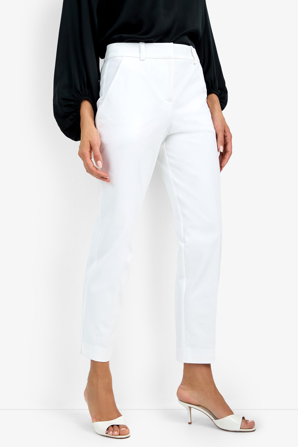 A woman in the Audrey Pant—white, tailored ankle-length trousers—pairs them with a black long-sleeve blouse and white open-toed heeled sandals, standing against a plain white background.