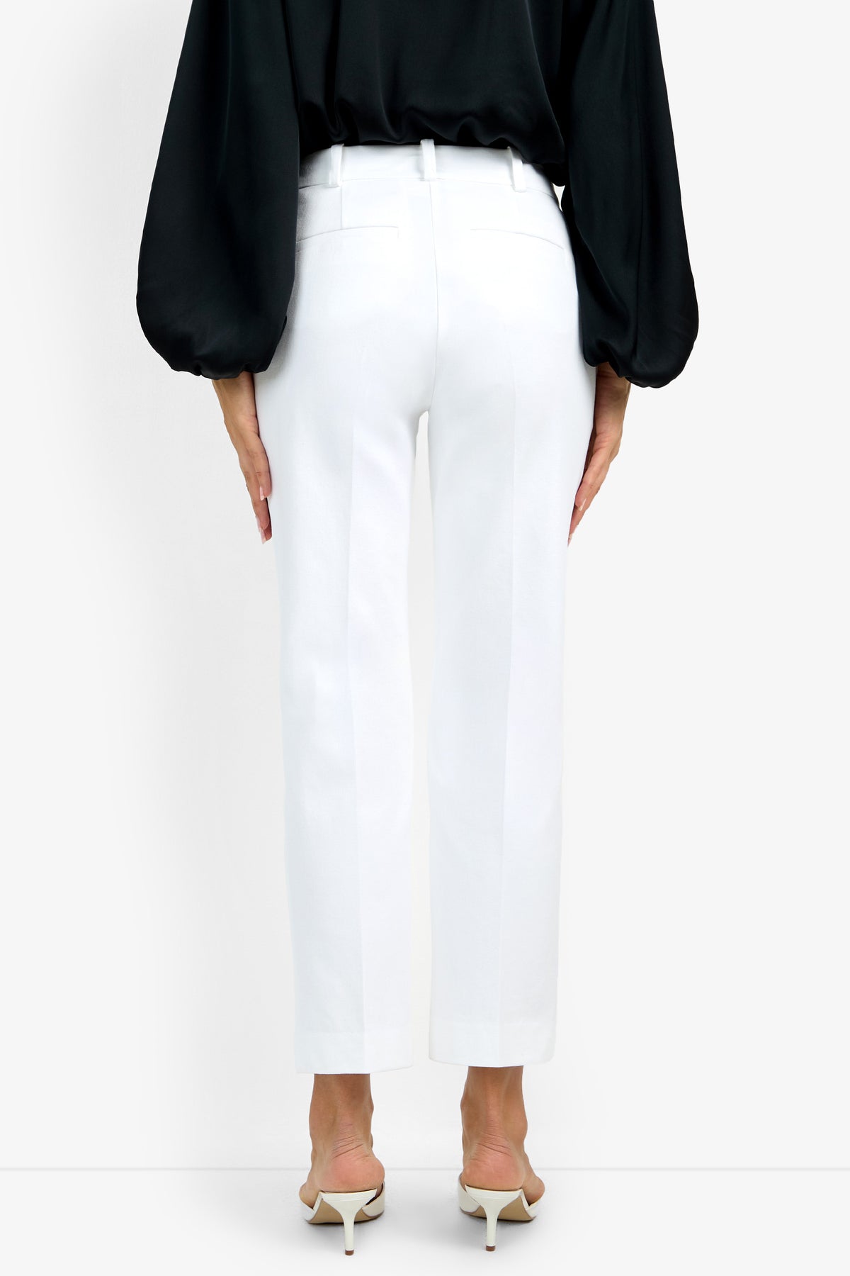 A person wearing the white, ankle-length Audrey Pant and a black long-sleeve blouse stands facing away, paired with white heeled sandals. The background is plain white.