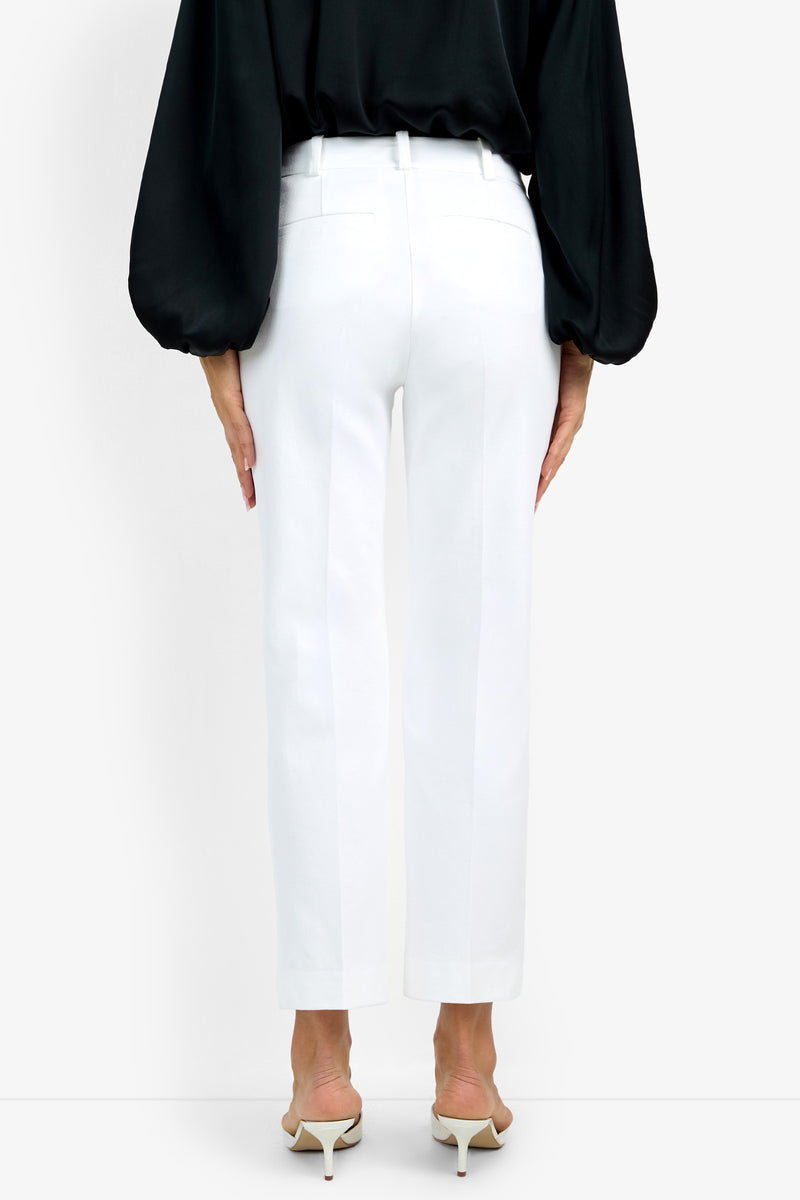 A person wearing the white, ankle-length Audrey Pant and a black long-sleeve blouse stands facing away, paired with white heeled sandals. The background is plain white.
