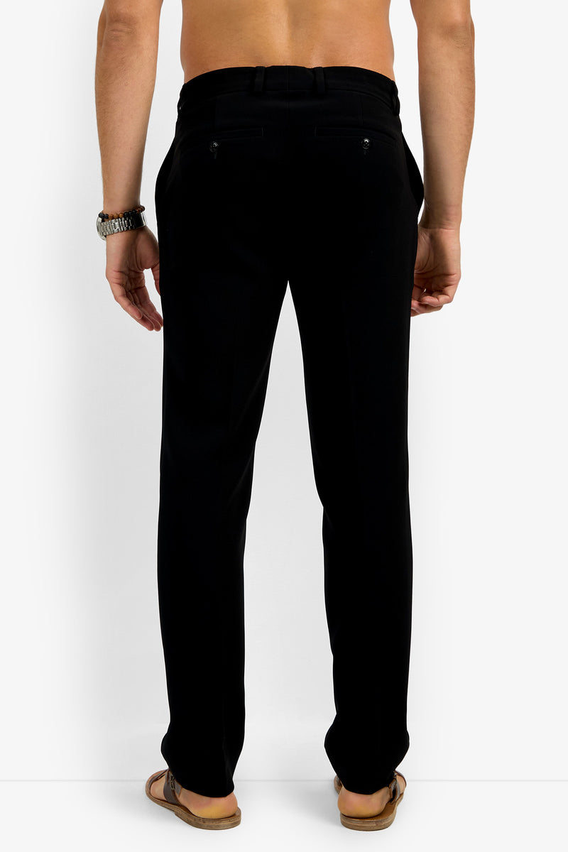 A man with his back to the camera wears Clyde Slim Trouser black dress pants, brown sandals, a wristwatch, and no shirt, set against a plain white background.