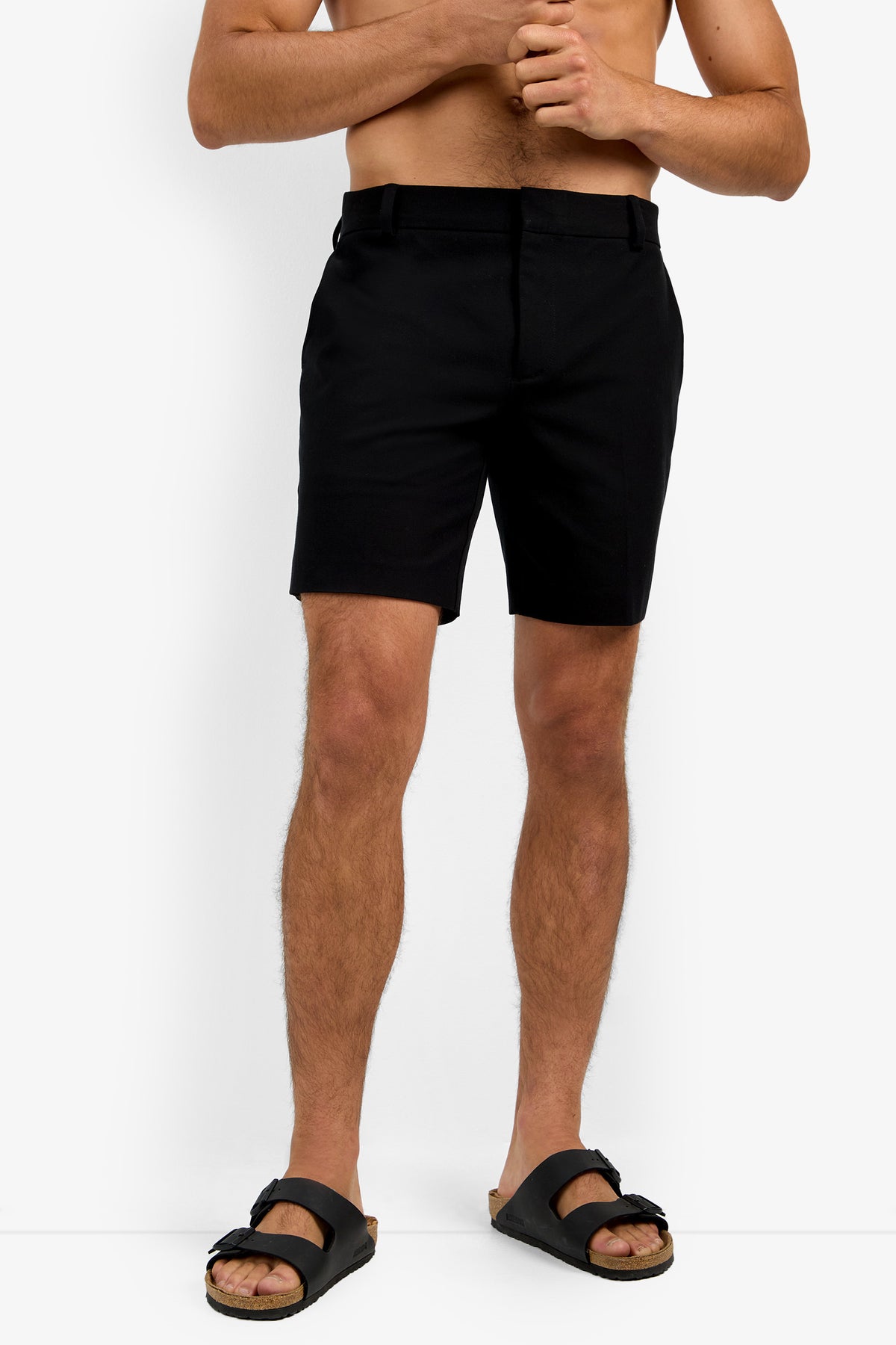 A shirtless person stands against a white background, wearing the Lawrence Short in black along with black sandals and hands clasped at the waist.
