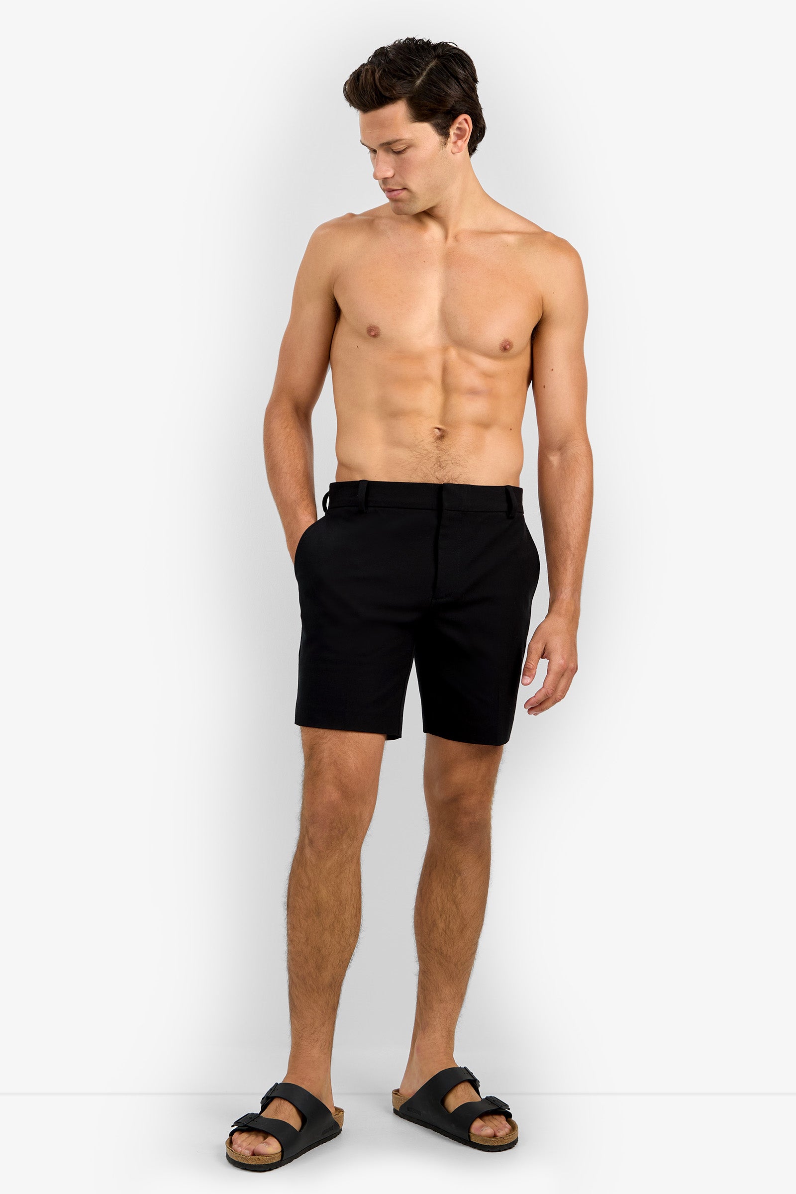 A shirtless man with short brown hair models the Lawrence Short in black, paired with black sandals. He stands against a plain white background, hands in his pockets, looking down.