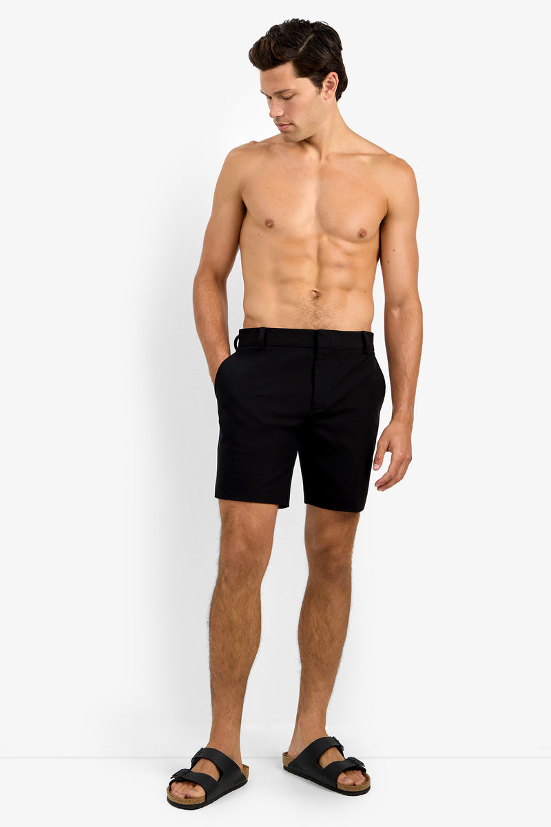 A shirtless man stands against a white background, wearing the Lawrence Short in black and black sandals. He has his left hand in his pocket and looks down toward his right side.