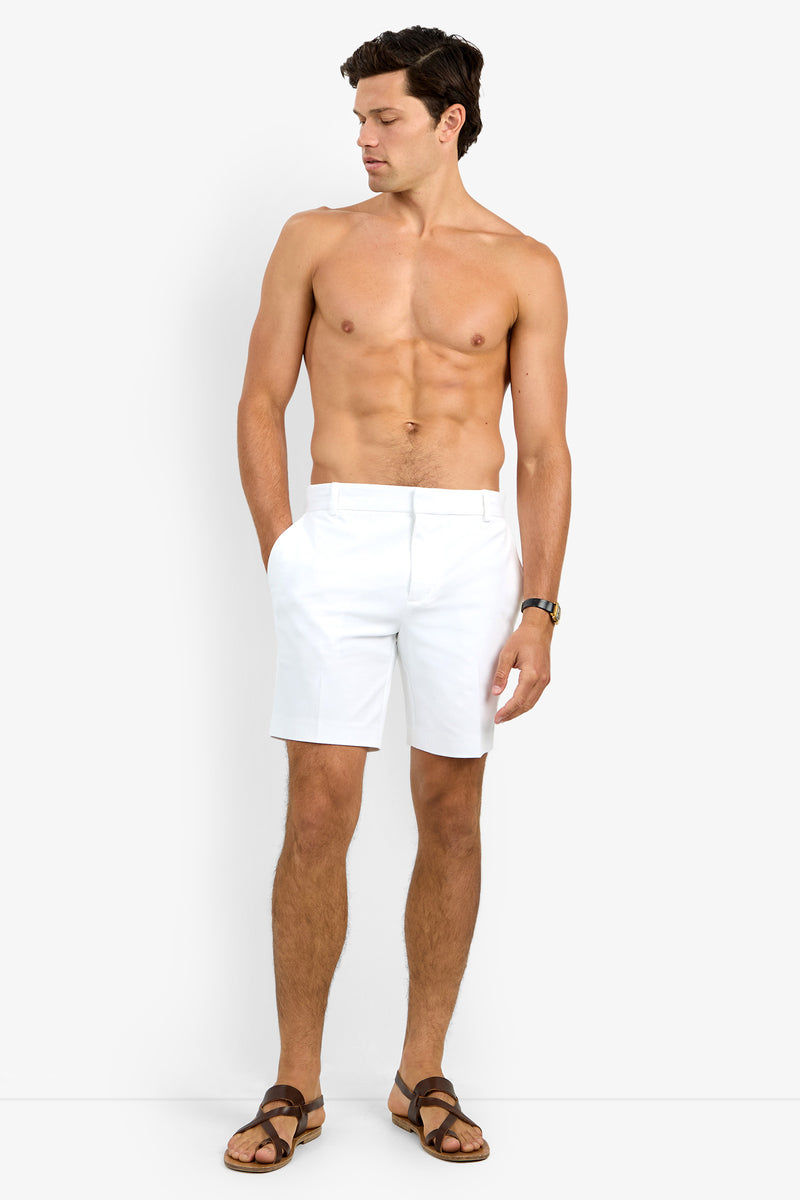 A man stands barefoot on a white background, wearing the Lawrence Short in white, brown sandals, and a wristwatch. He is shirtless, looking down to his left with his hands in his pockets.