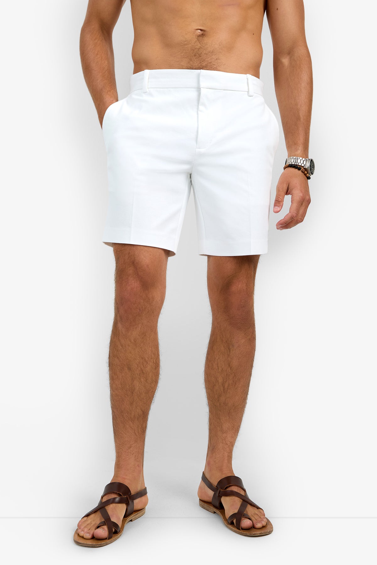 A man models the Lawrence Short in white, paired with brown sandals and a silver watch, standing against a plain white background. His torso is bare and his hands are in his pockets.