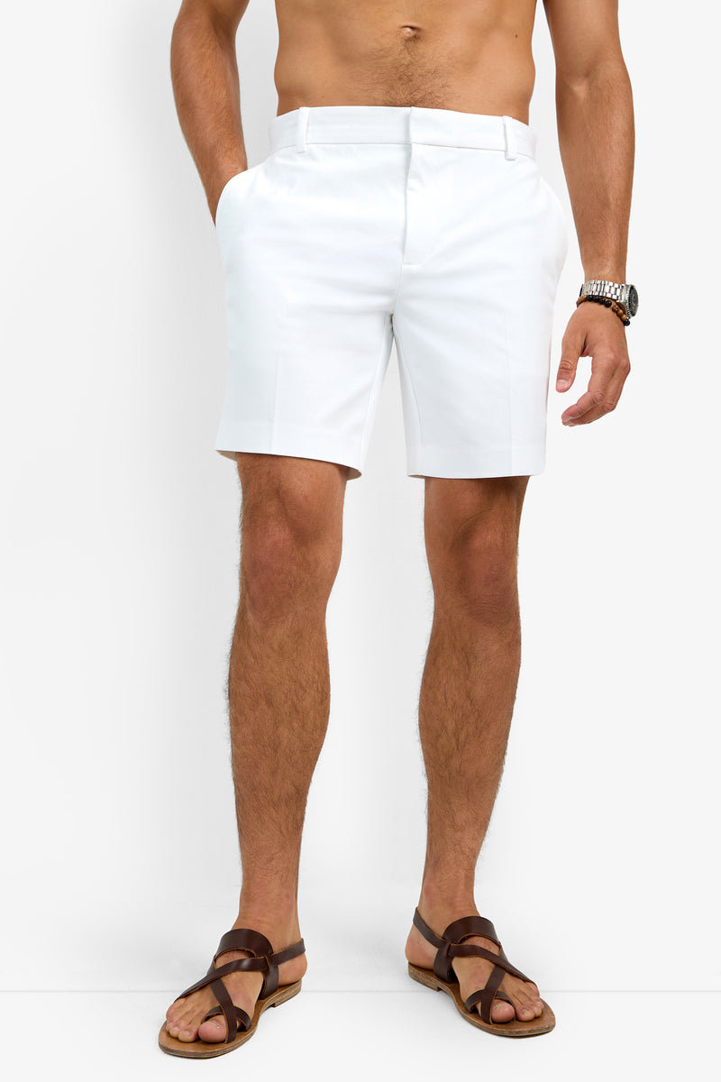 A shirtless man stands against a plain white background, wearing the Lawrence Short in white, brown sandals, and a wristwatch with his hands in his pockets.