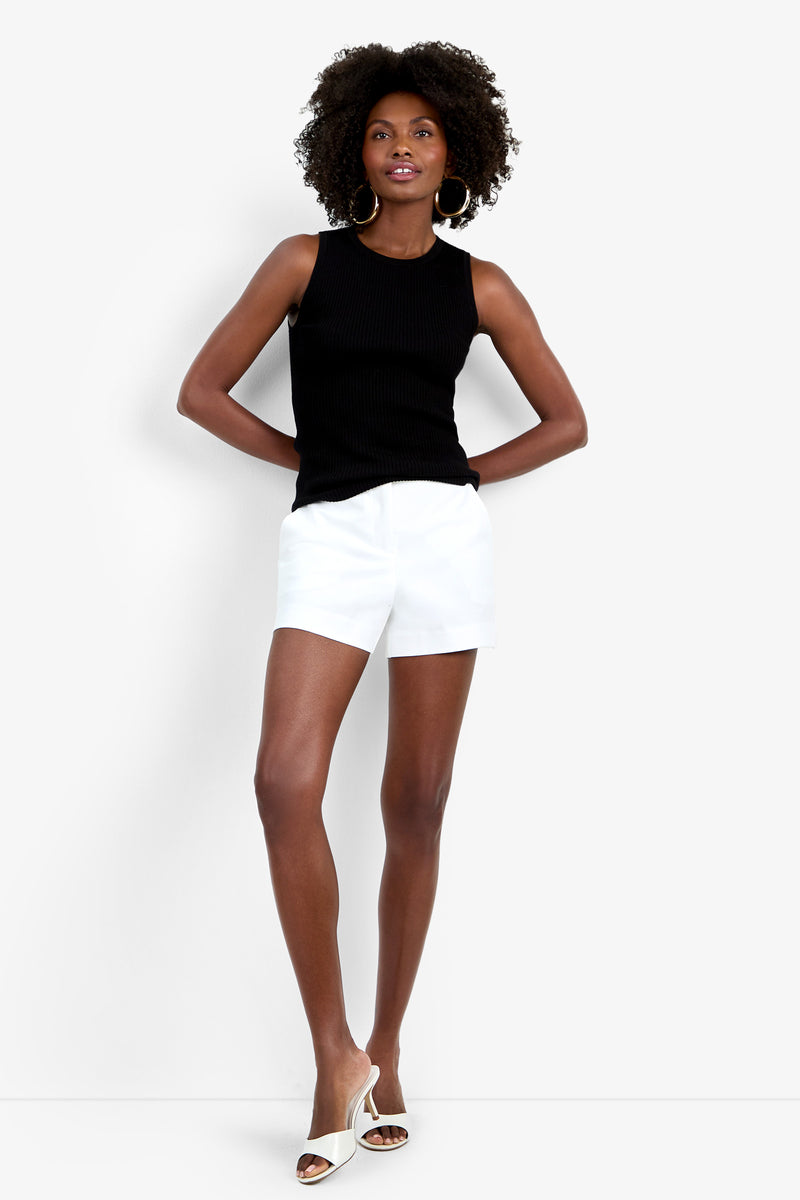 A woman with curly hair poses confidently against a white background wearing the sleeveless black Hampton Crewneck Top, white shorts, white heels, and large hoop earrings, with one hand on her hip.