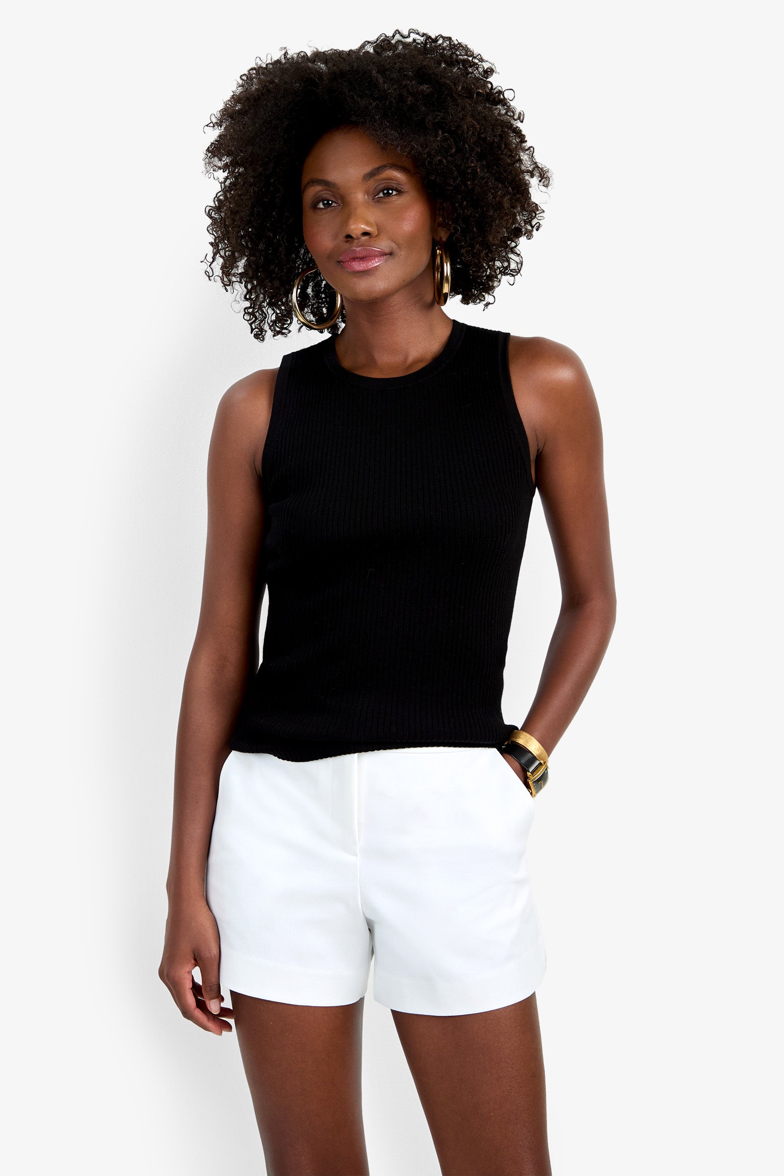 A woman with curly hair smiles softly against a plain white background, wearing the sleeveless black Hampton Crewneck Top, white shorts, gold hoop earrings, and a gold bracelet.