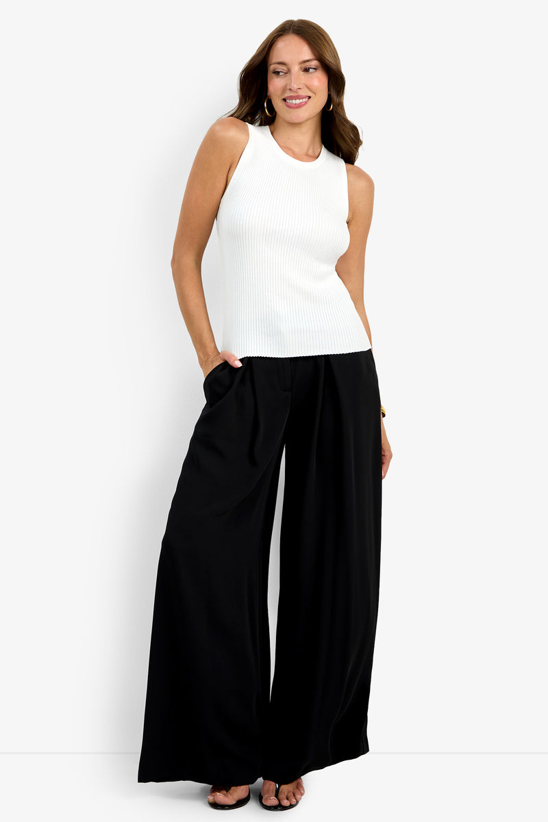 A woman smiles as she poses against a plain white background, wearing the sleeveless white Hampton Crewneck Top and high-waisted, wide-leg black pants with her hands in the pockets.
