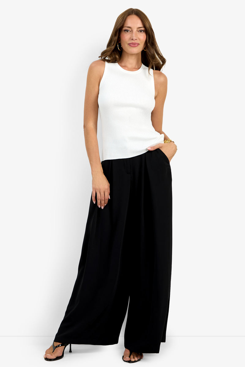A woman stands against a white background, wearing the Hampton Crewneck Top (sleeveless, white, ribbed) paired with loose black wide-leg pants and black sandals. She poses with one hand in her pocket and smiles slightly.