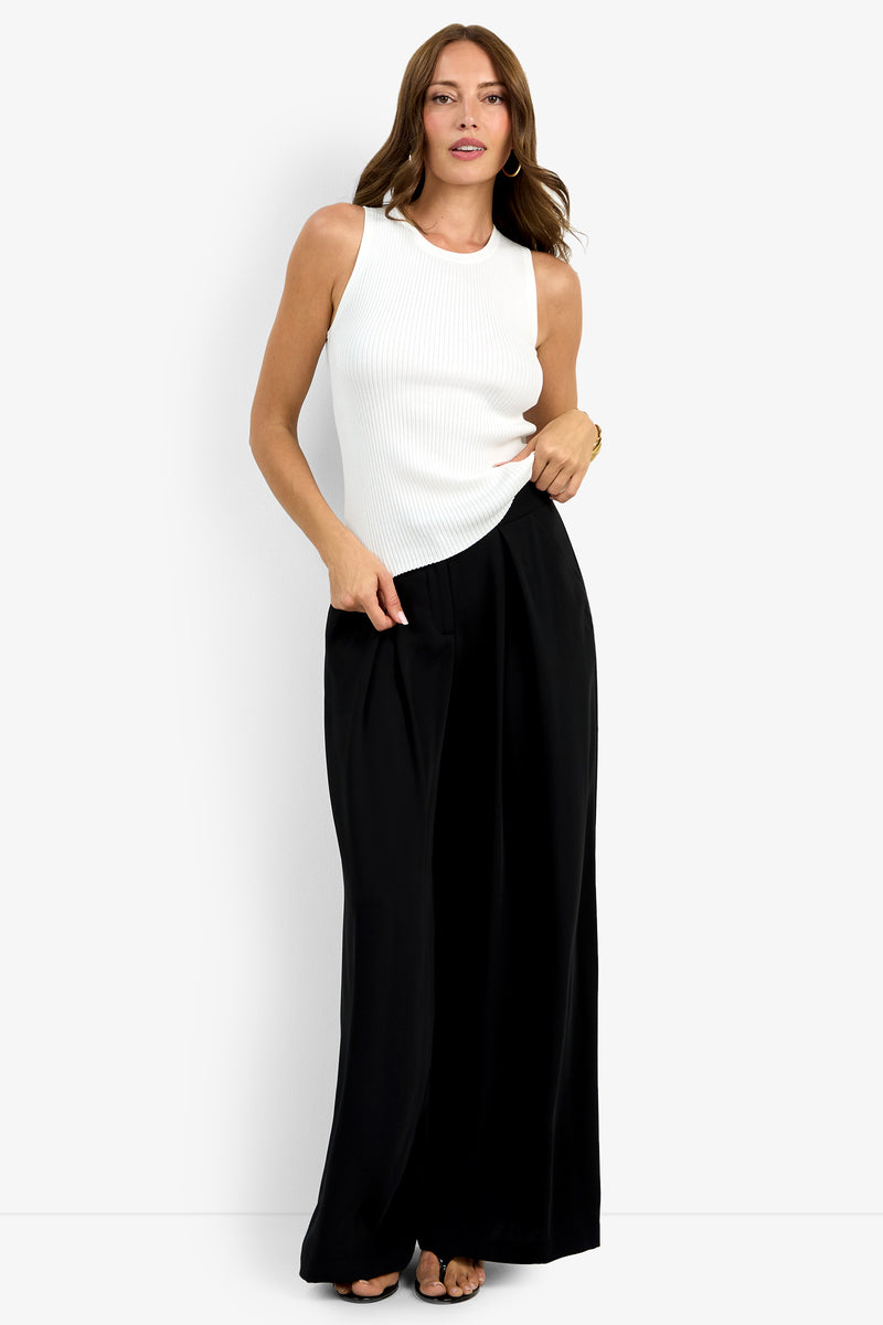 A woman with long brown hair poses confidently against a white background, wearing the sleeveless, fitted Hampton Crewneck Top in white and high-waisted, wide-leg black pants with open-toe shoes.