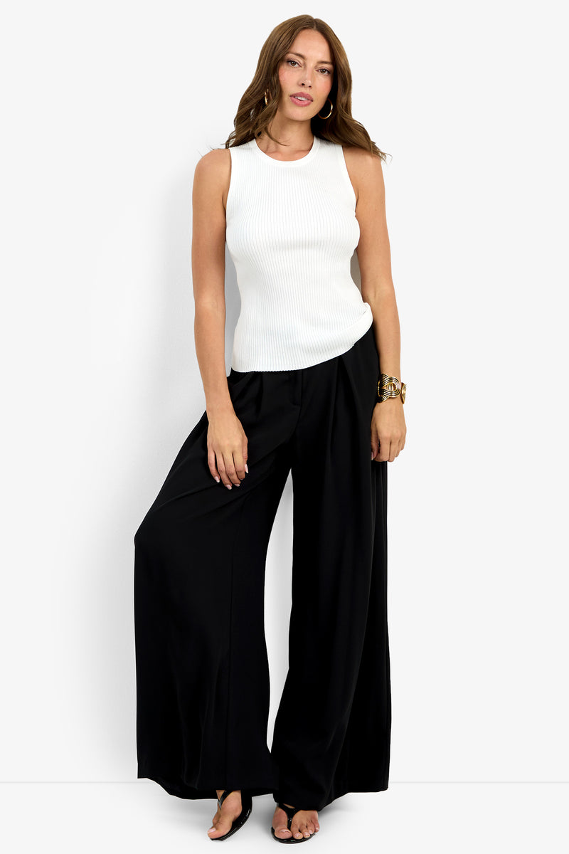 A woman with long brown hair stands confidently against a white background wearing the Hampton Crewneck Top, loose black wide-leg pants, black sandals, and a chunky bracelet, posed with one leg crossed.