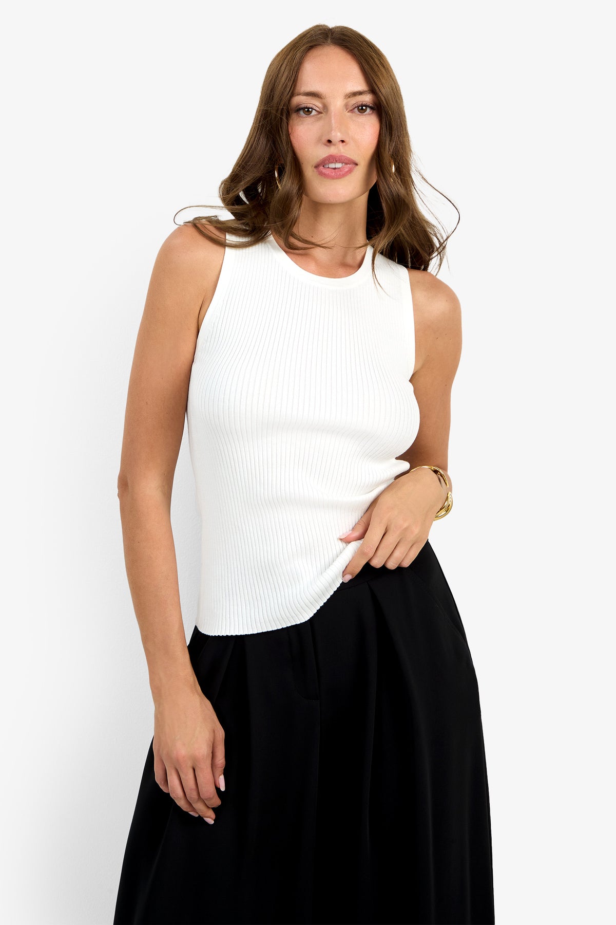 A woman with long brown hair wears the Hampton Crewneck Top, sleeveless and white, paired with a black skirt. She stands against a plain white background, looking at the camera with one hand on her hip.