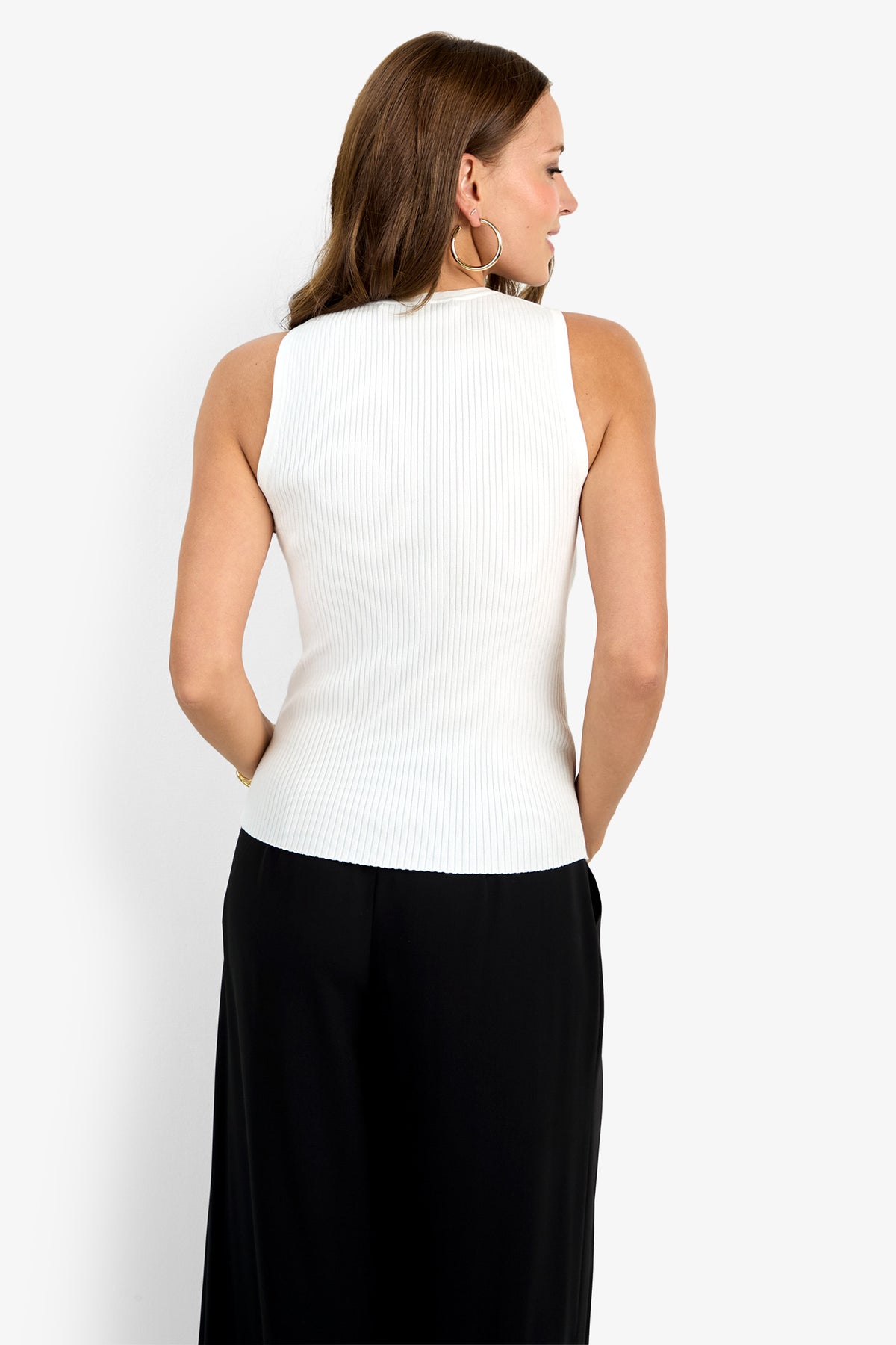 A woman with long brown hair, wearing large hoop earrings and the white ribbed sleeveless Hampton Crewneck Top with black pants, stands facing away from the camera against a plain white background.