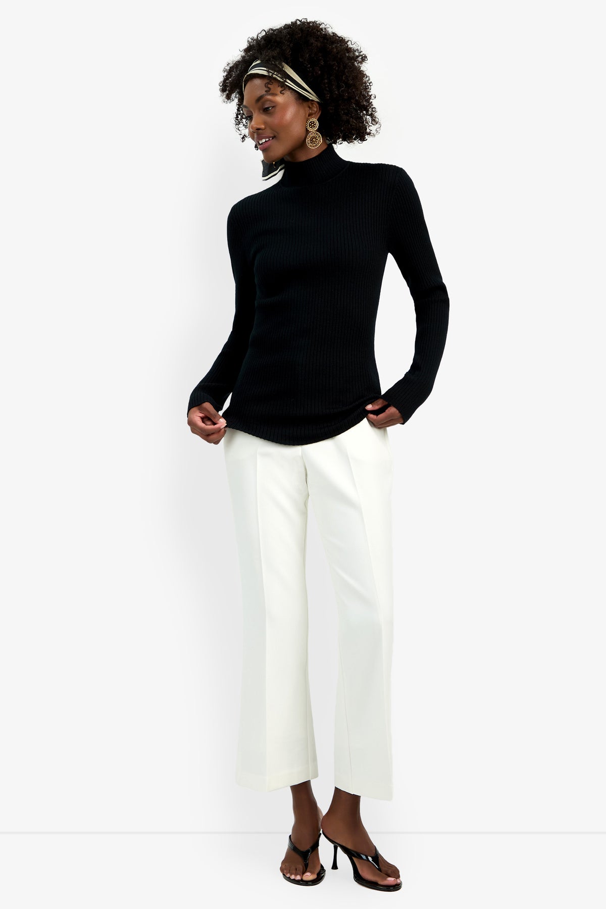 A woman wearing the Hempstead Mock Neck Top, white wide-leg trousers, heeled sandals, large hoop earrings, and a patterned headband stands smiling and looking to the side.