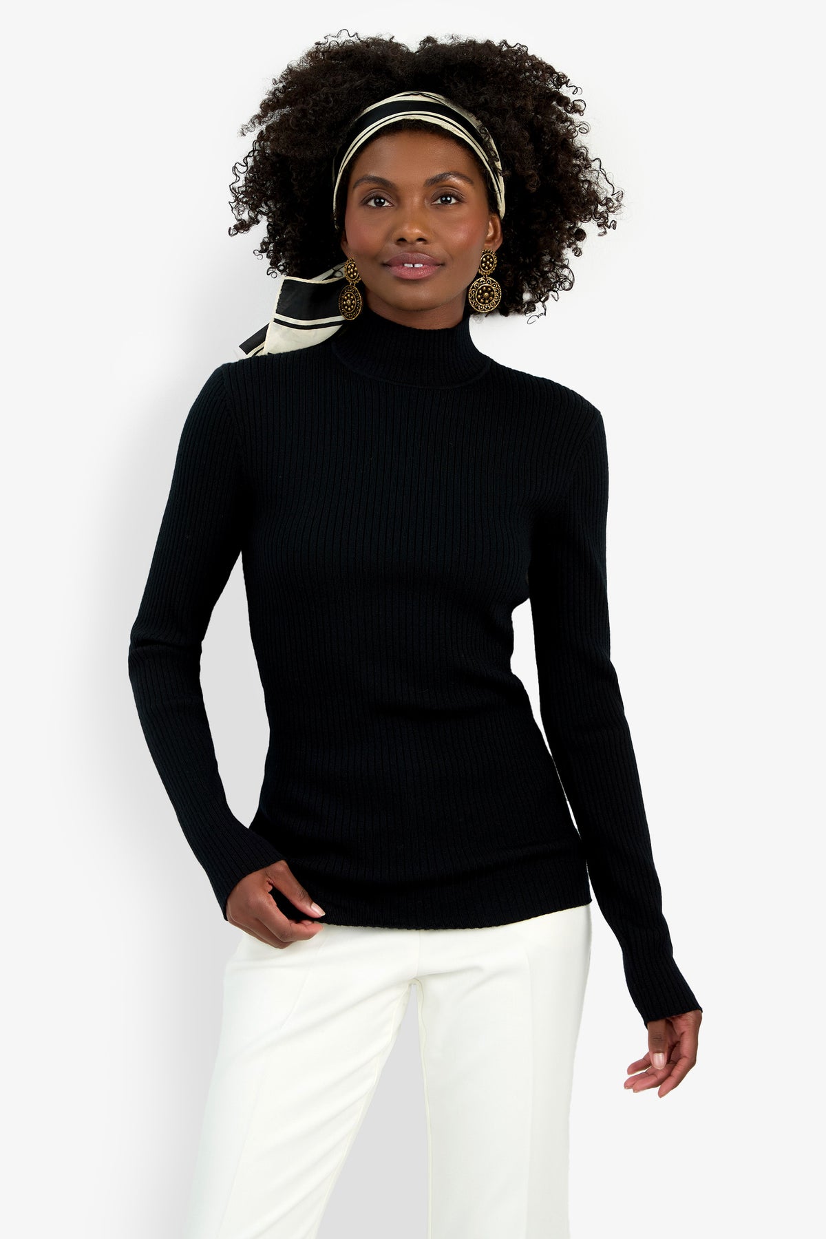 A woman with curly hair wears the Hempstead Mock Neck Top in black, white pants, gold earrings, and a black-and-white striped headband while standing against a plain white background.