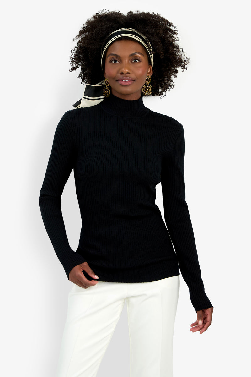 A woman with curly hair wears the Hempstead Mock Neck Top in black, white pants, gold earrings, and a black-and-white striped headband while standing against a plain white background.