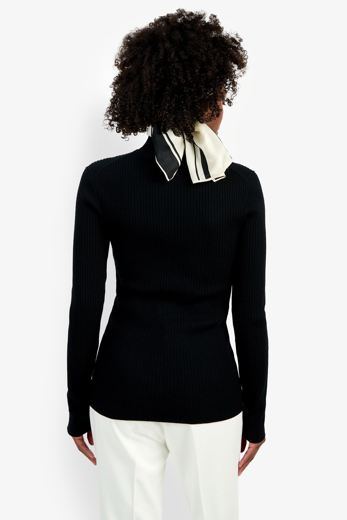 Wearing the Hempstead Mock Neck Top in black, paired with white pants and a black-and-white neck scarf, a curly-haired person stands with their back to the camera against a plain white background.
