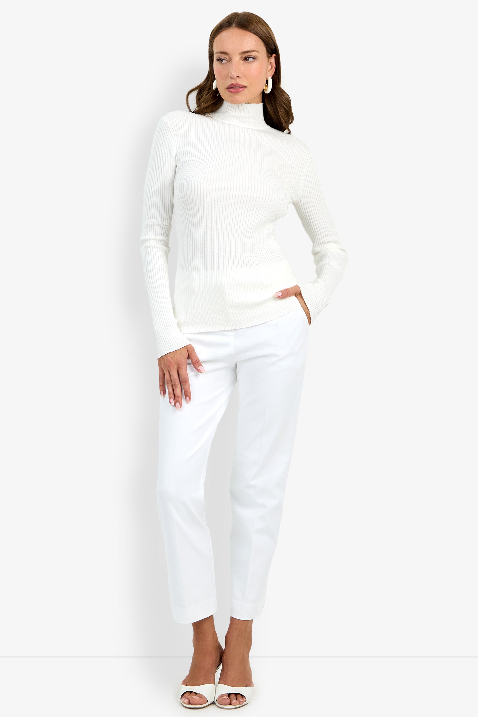 A woman stands against a white background wearing the Hempstead Mock Neck Top, white pants, and white open-toe heels. One hand is in her pocket as she gazes to the side with a neutral expression.