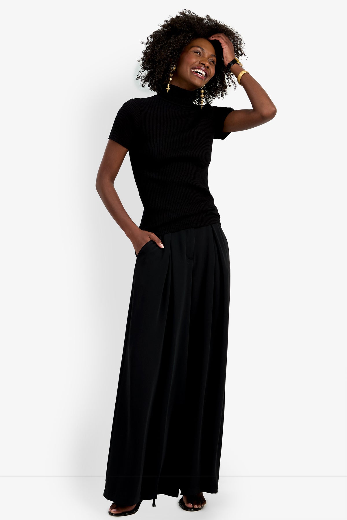 A smiling woman with curly hair models the Armeni Mock Neck Top, a short-sleeve black turtleneck, paired with a long black pleated skirt. She stands against a white background, one hand in her pocket and the other touching her hair.