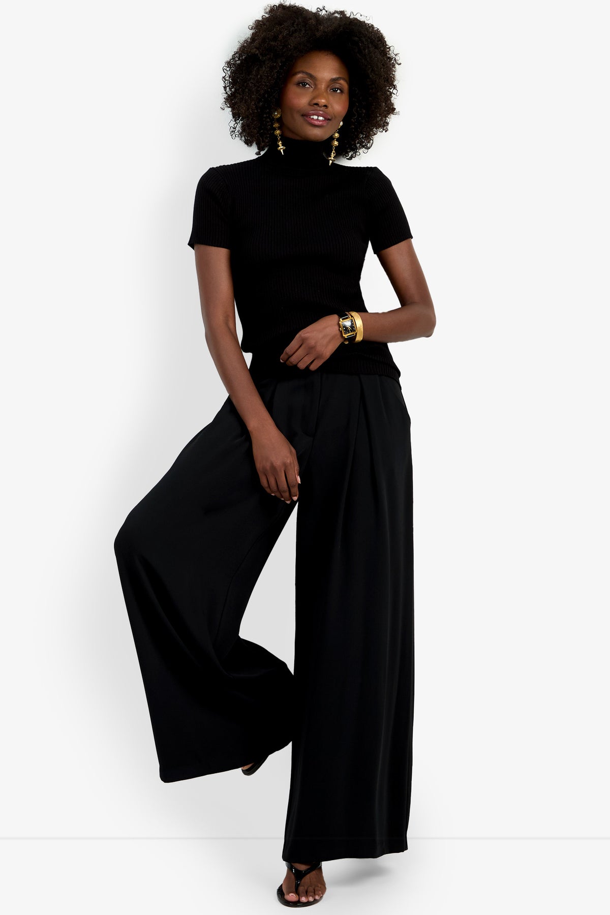 A woman with curly hair poses against a white background, wearing the Armeni Mock Neck Top in black with wide-leg black pants, gold earrings, a gold bracelet, and black sandals; she lifts one leg.