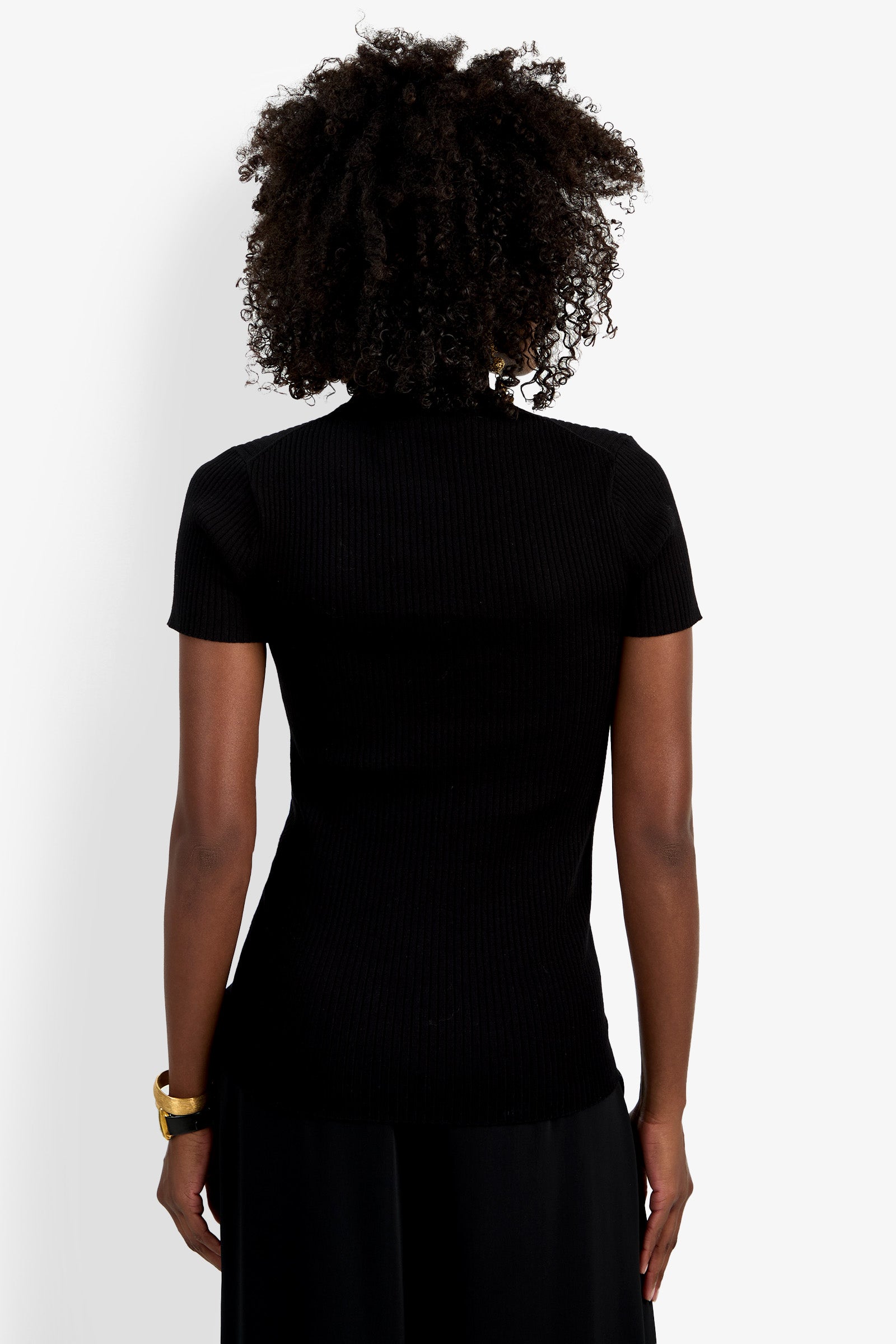 A person with curly hair, wearing the Armeni Mock Neck Top and a gold bracelet, stands facing away from the camera against a plain white background.
