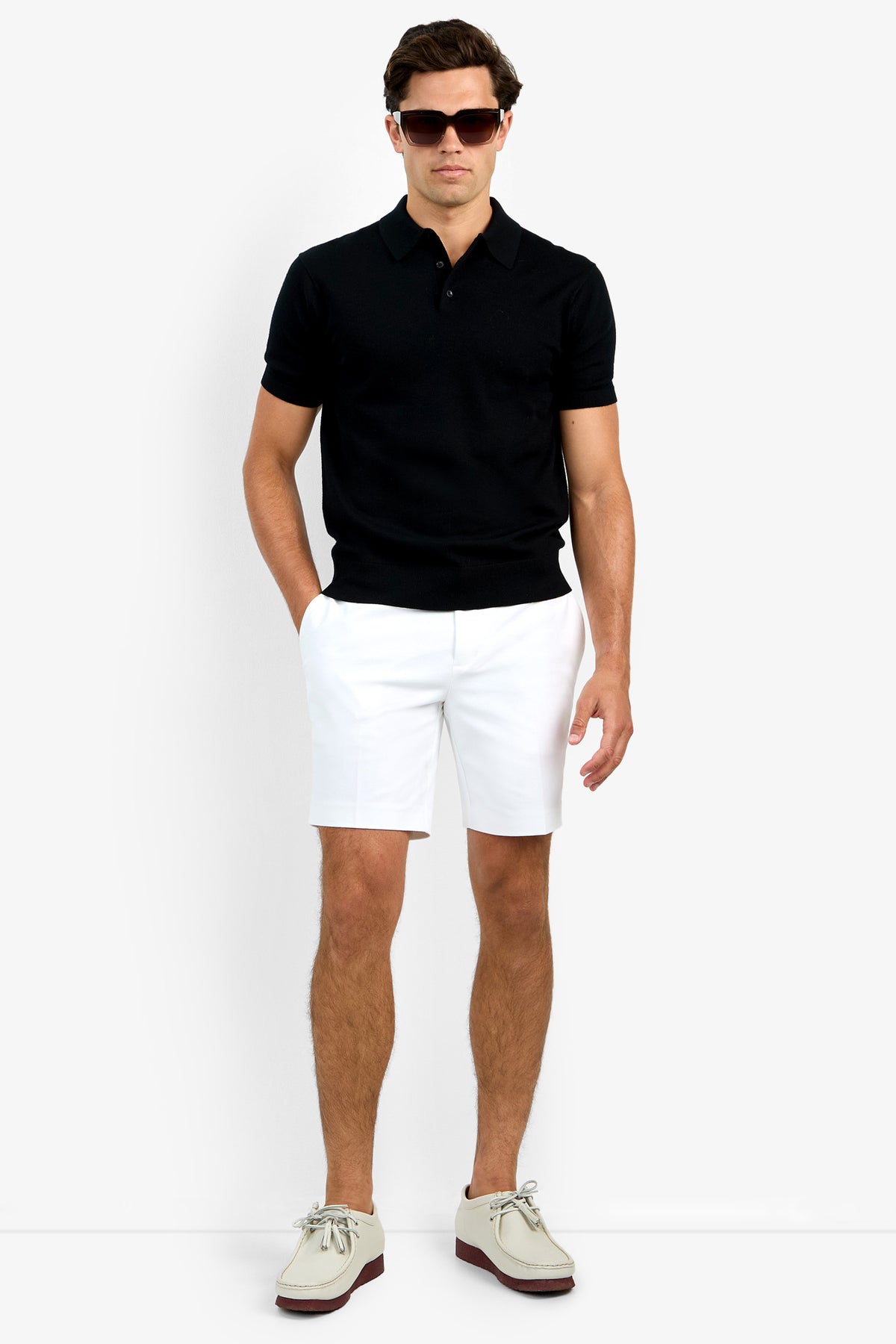 A man wearing the Grant Short Sleeve Polo, white shorts, beige boat shoes, and sunglasses stands with his hands in his pockets against a plain white background.
