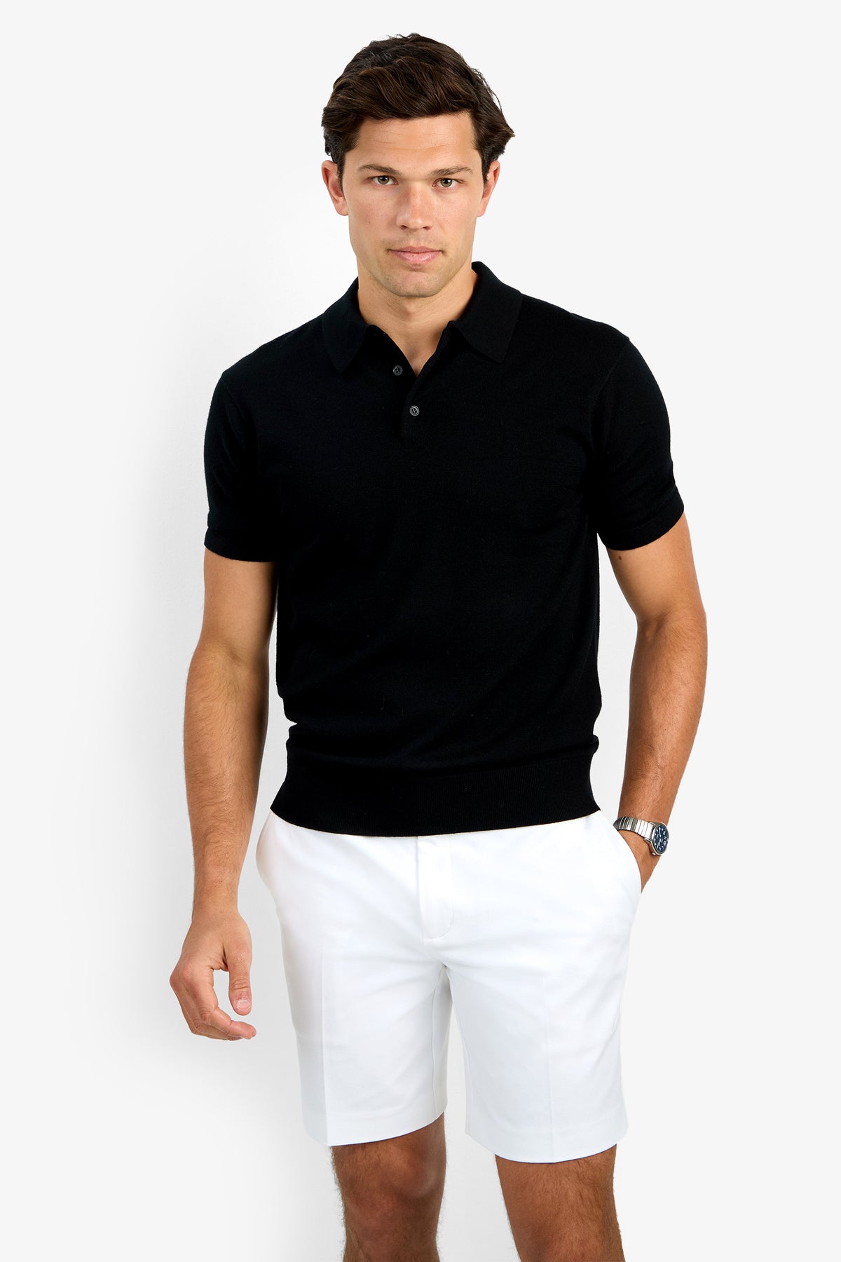 A man with short brown hair and a neutral expression stands against a plain white background, wearing the Grant Short Sleeve Polo in black, paired with white shorts and a watch on his left wrist.