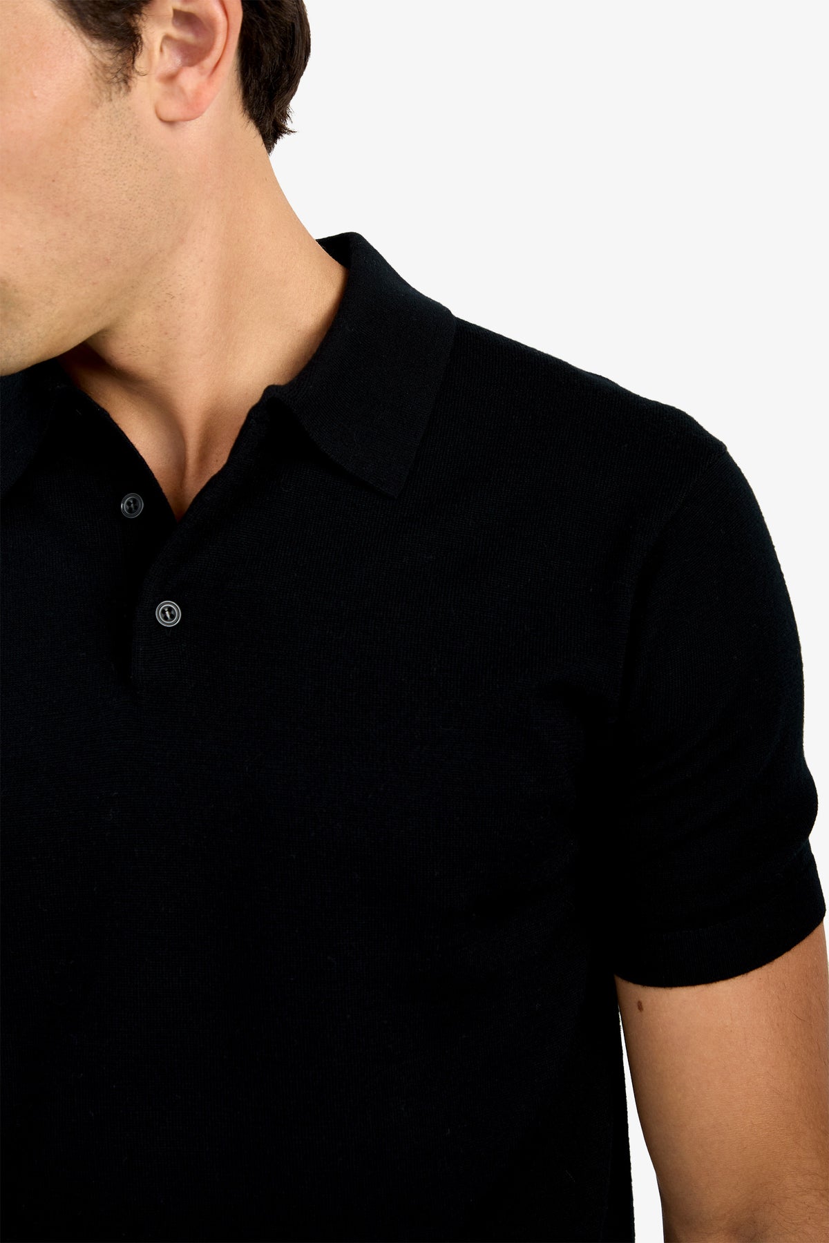 A man models the Grant Short Sleeve Polo in black, shown from shoulders to mid-arm, facing slightly left with his face partially out of frame against a white background.