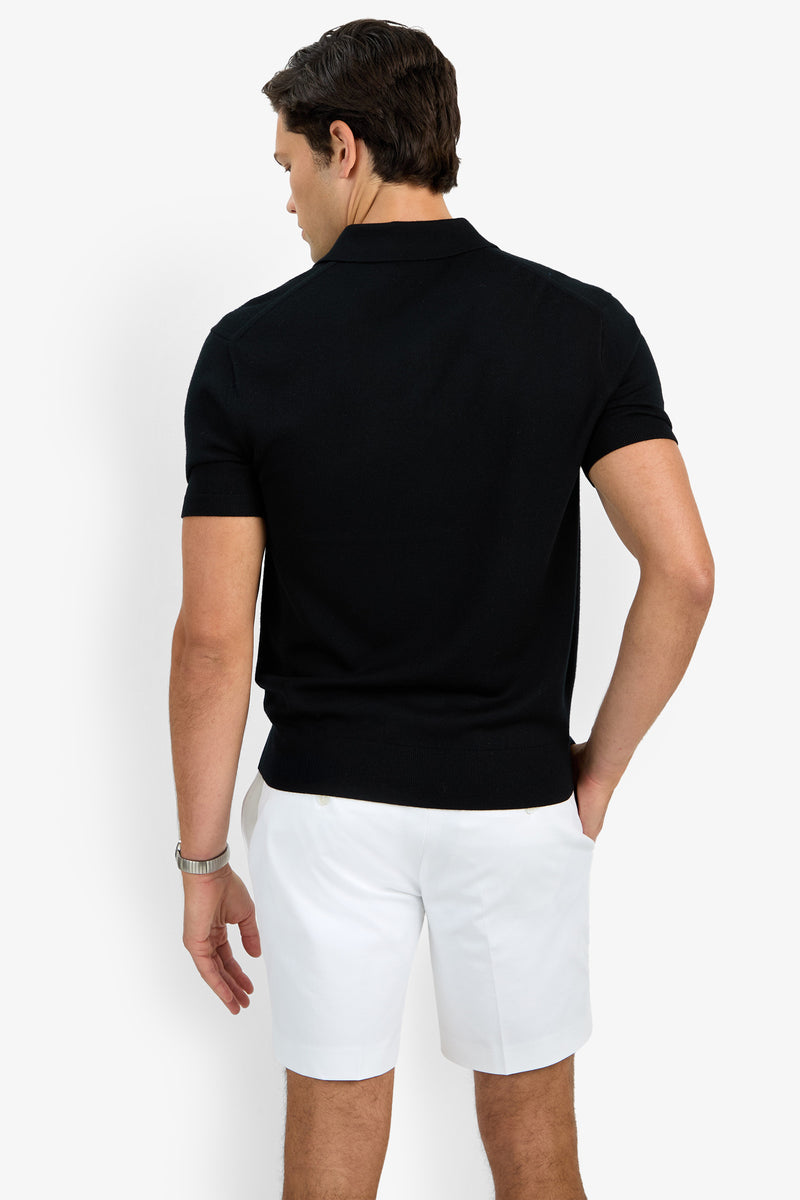 A man stands with his back to the camera wearing the black Grant Short Sleeve Polo and white shorts, one hand in his pocket and a watch on his wrist. The background is plain white.