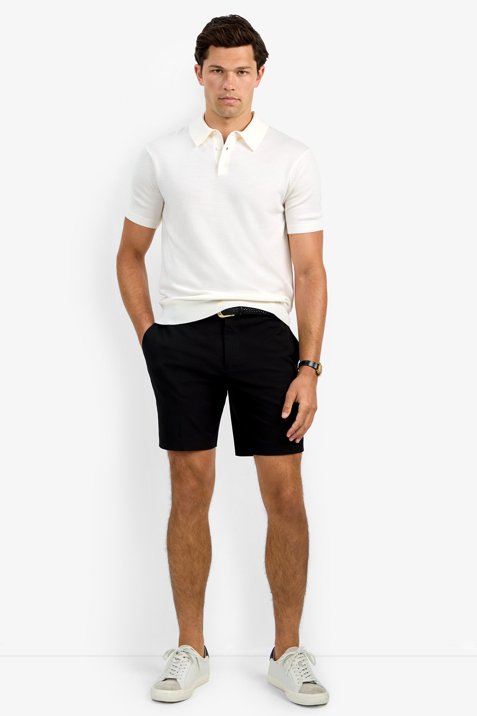 A man with short brown hair stands against a plain background wearing the Grant Short Sleeve Polo, black shorts, white sneakers, and a black belt. He has a neutral expression and his hands are in his pockets.