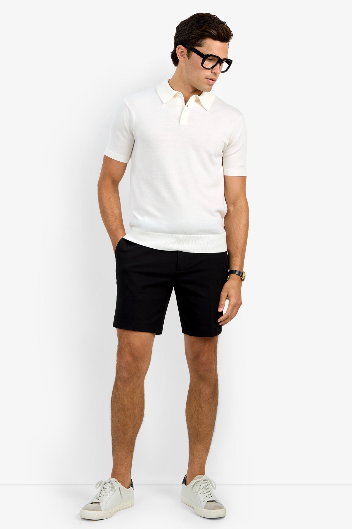 A man wears the Grant Short Sleeve Polo, black shorts, white sneakers, a wristwatch, and black-rimmed glasses, standing against a plain light background with his hands in his pockets, looking down to his right.