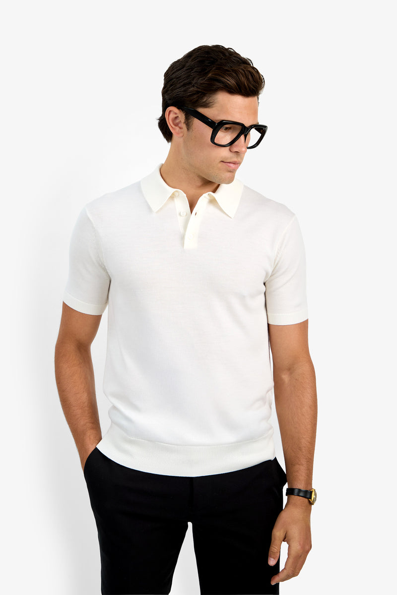 A man wearing the Grant Short Sleeve Polo, black pants, thick black glasses, and a watch stands with one hand in his pocket, looking down and to the side against a plain white background.