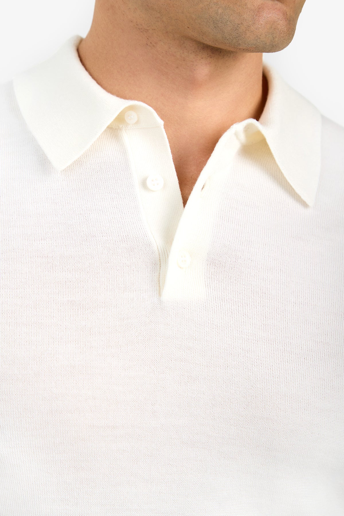 Close-up of a person wearing the Grant Short Sleeve Polo, an off-white knit shirt with a collar and three buttons, showing the upper chest and neck. The background is plain and light-colored.